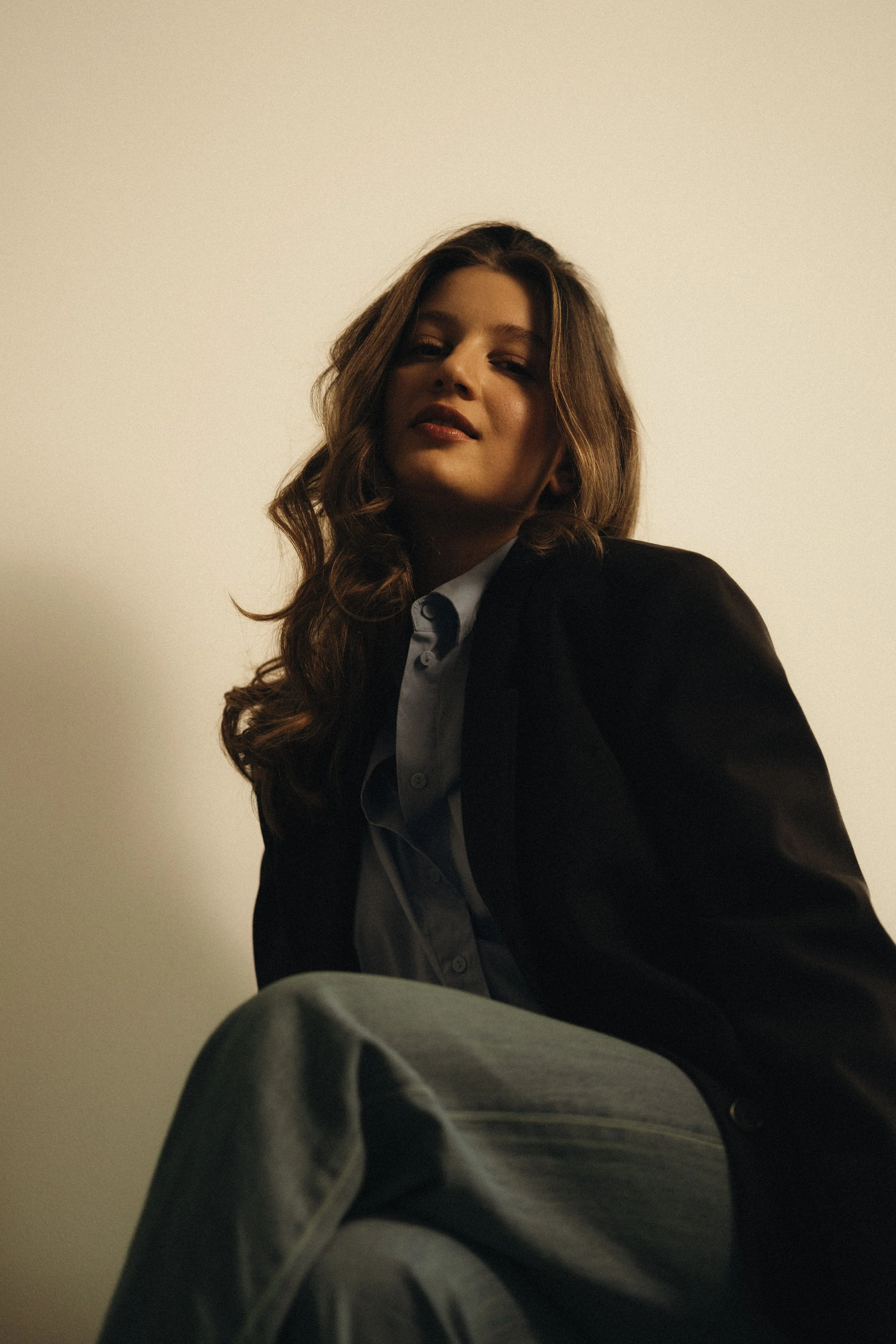 Porträt einer jungen Frau mit lang, gewelltem Haar, die einen schwarzen Blazer und eine hellblaue Bluse trägt, sitzt vor einem hellen Hintergrund und blickt selbstbewusst in die Kamera.