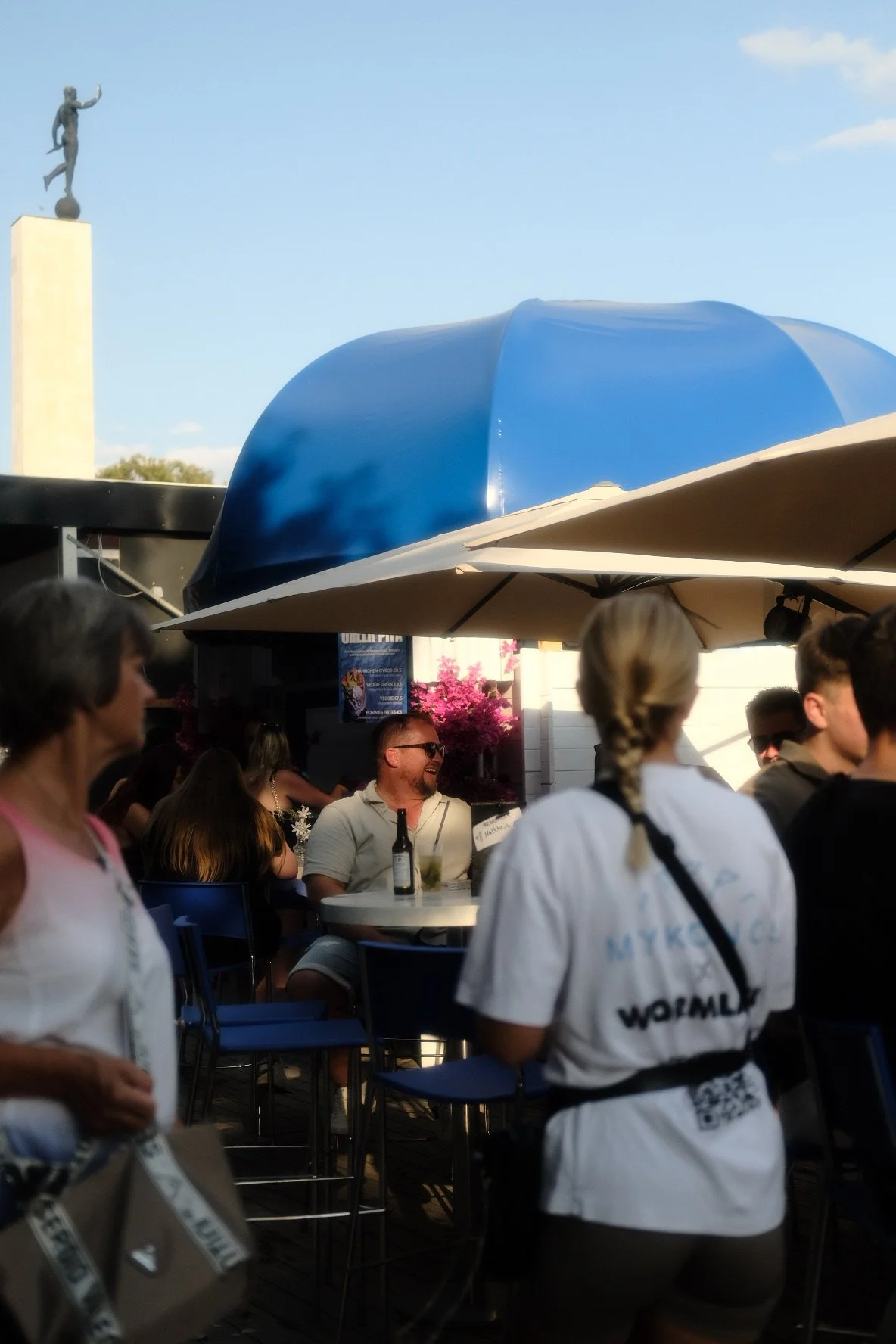 Menschen sitzen im Biergarten, einige lachen und sprechen, mit einigen Getränken auf Tischen, im Hintergrund blühende Bäume, Sonnenschirme und eine Statue im Freien.