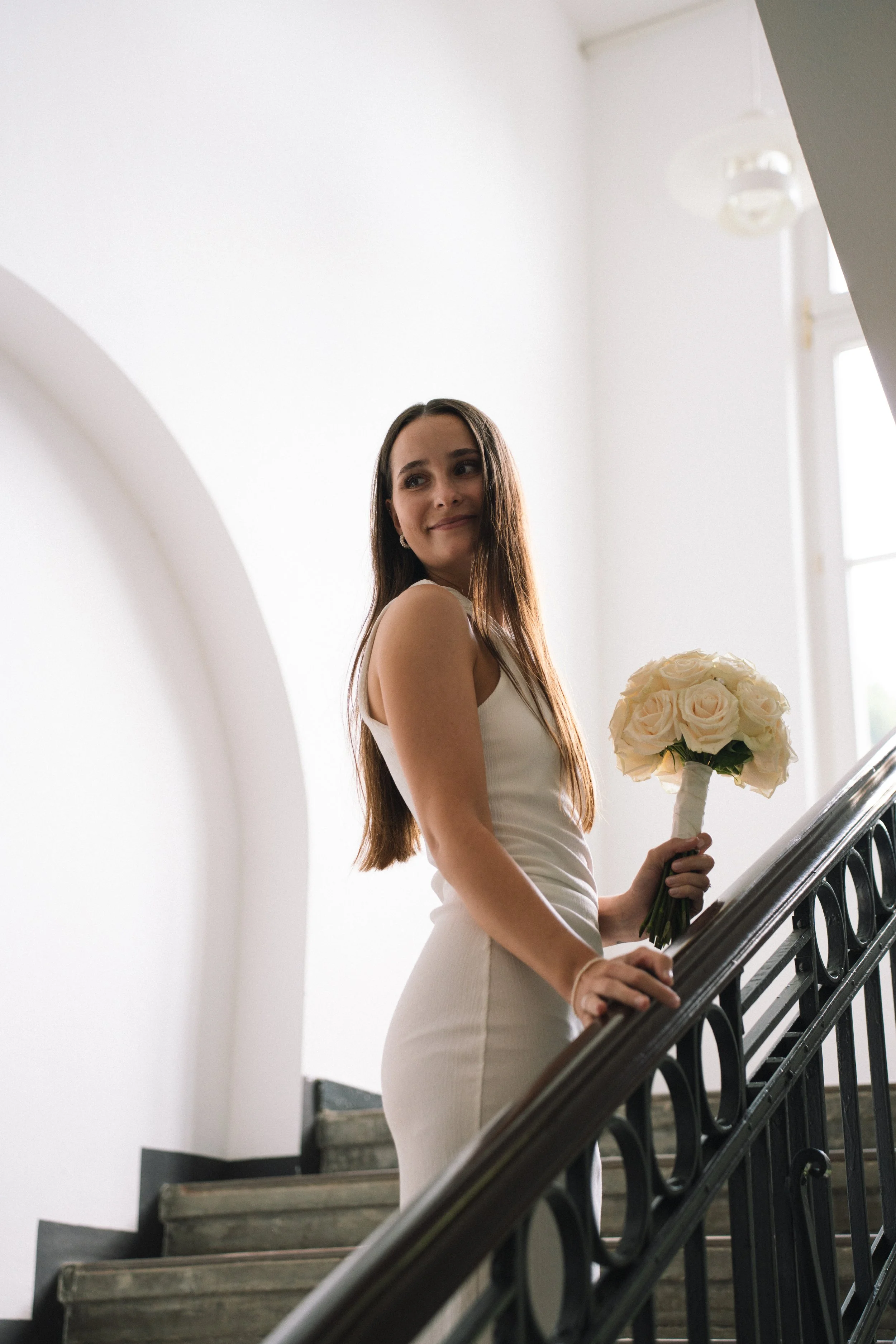 Junge Frau in einem weißen Kleid steht auf einer Treppe im Innenbereich mit weißem Hintergrund und hält einen Blumenstrauß. Sie schaut nach rechts und lächelt leicht.