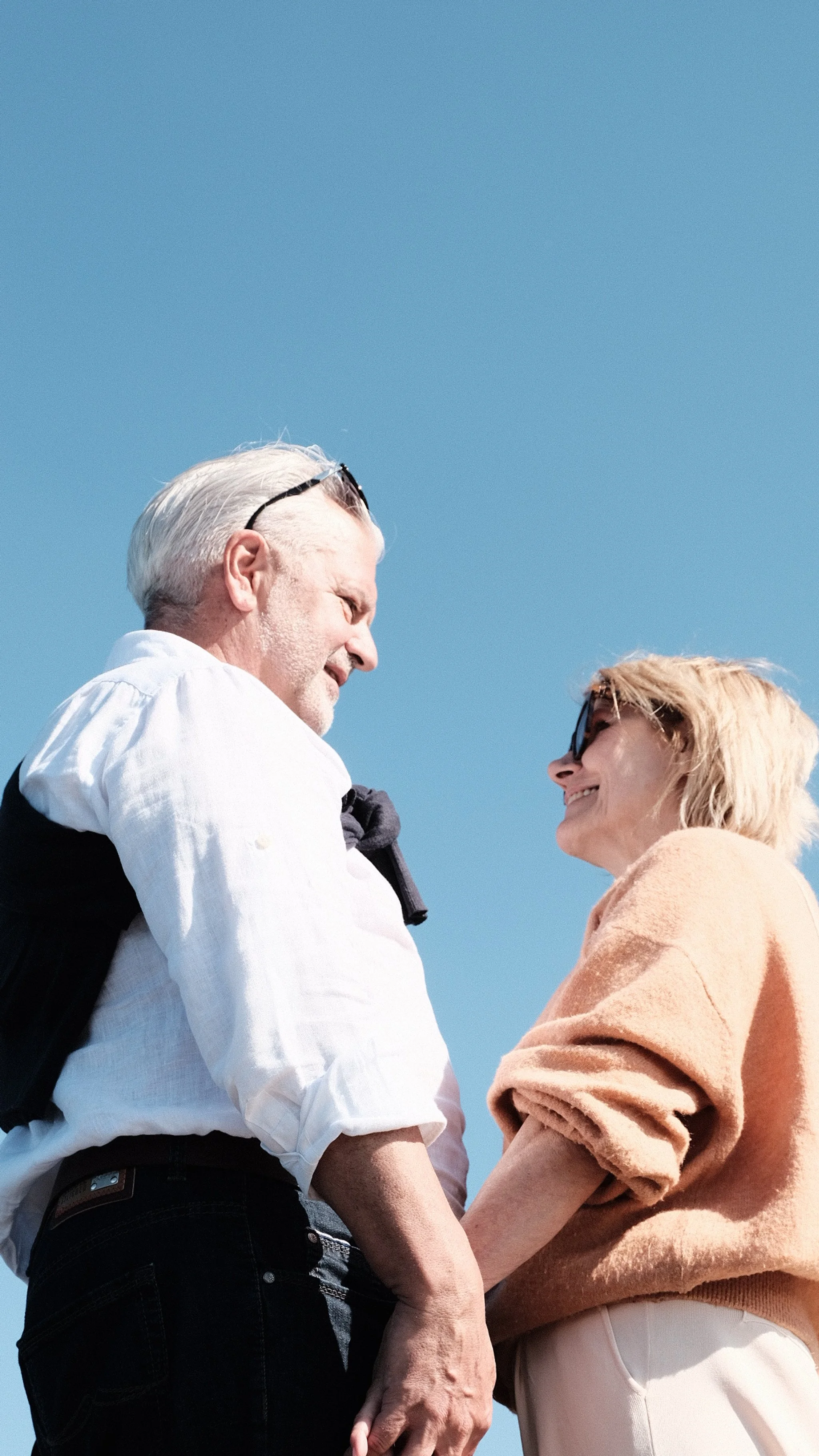 Ein älteres Paar, ein Mann mit weißen Haaren und eine Frau mit blonden Haaren, schaut sich liebevoll an und hält Händchen bei schönem Wetter mit blauem Himmel.