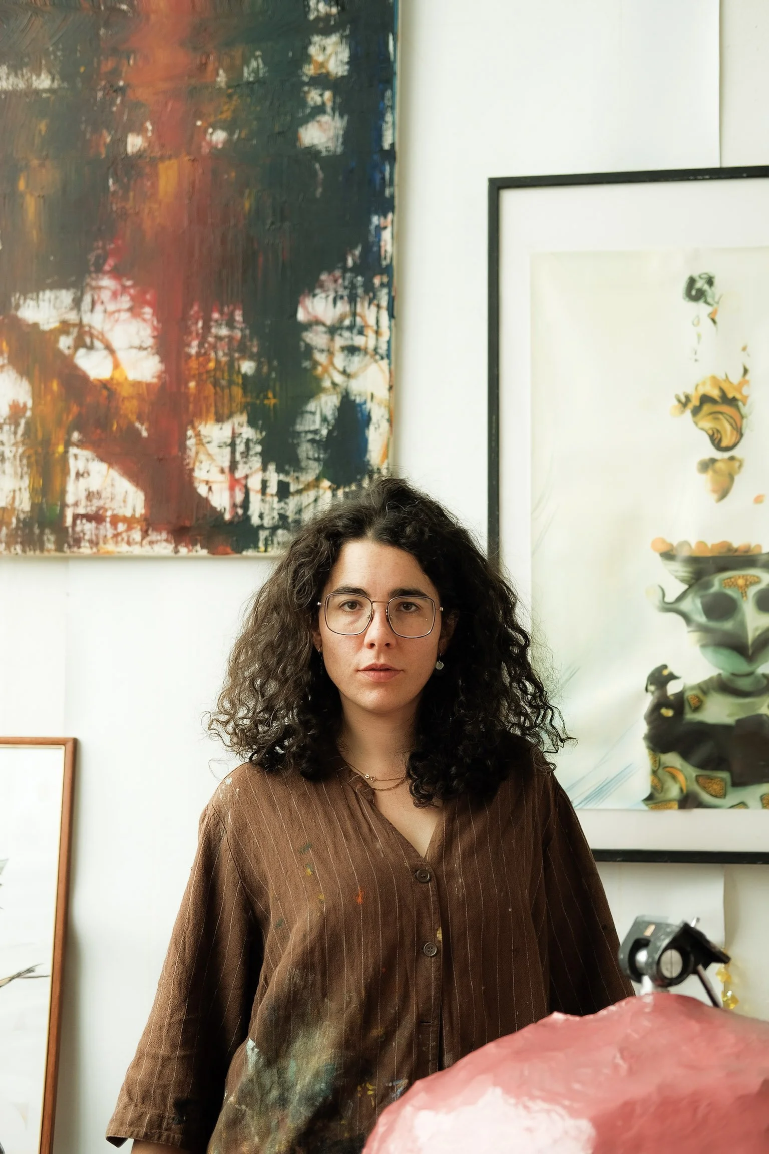 Junge Frau mit dunklen, lockigen Haaren und Brille in einem Kunststudio, umgeben von abstrakten Gemälden an der Wand.