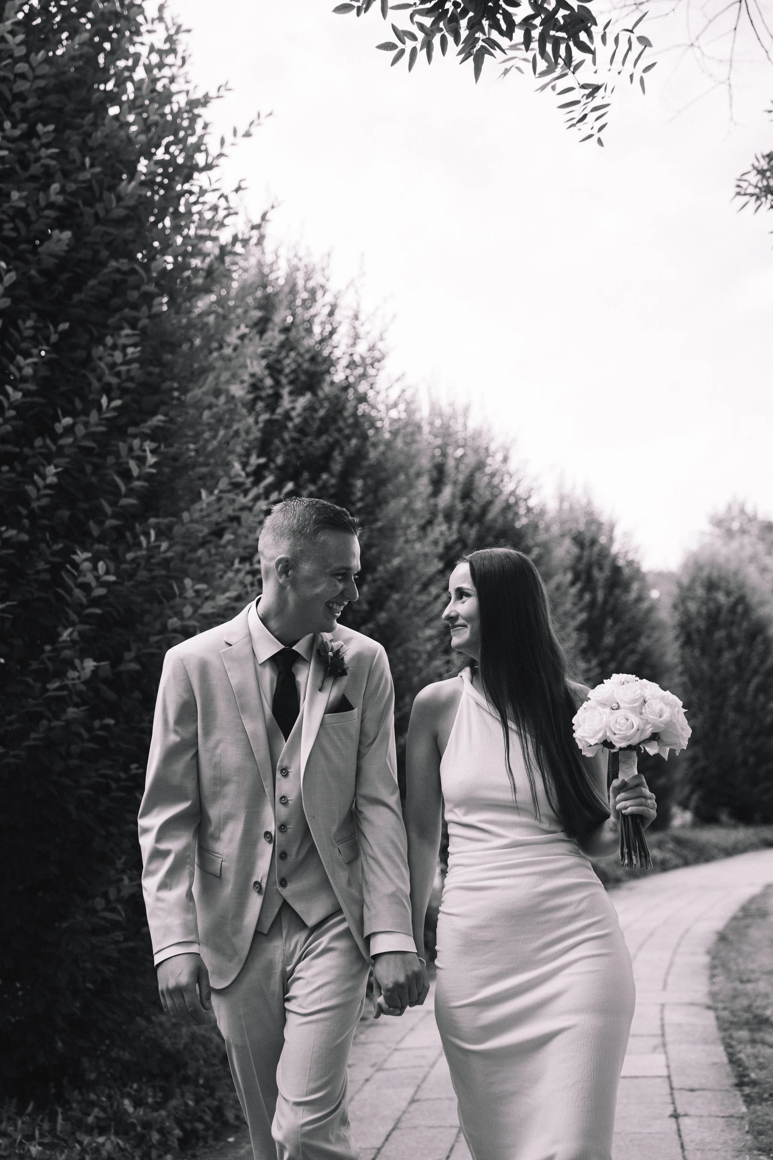 Ein lachendes Paar in Hochzeitskleidung geht Hand in Hand auf einem Weg im Freien, umgeben von Bäumen, bei Sonnenuntergang. Sie tragen elegante Kleidung, er einen Anzug und sie ein weißes Kleid mit einem Blumenstrauß.