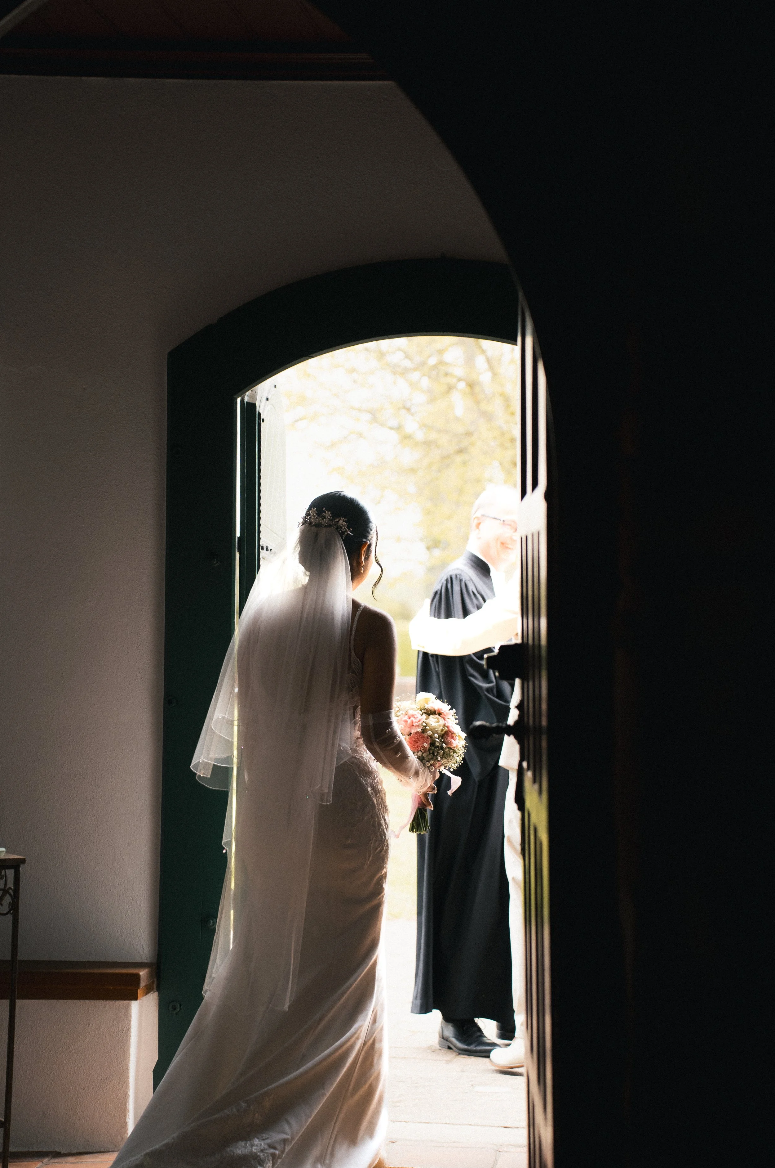 Braut in einem weißen Kleid mit Schleier und Blumenstrauß steht an der Tür eines dunklen Raumes, während ein gutaussehender Mann in dunkler Robe den Raum betritt, bei Tageslicht im Freien.