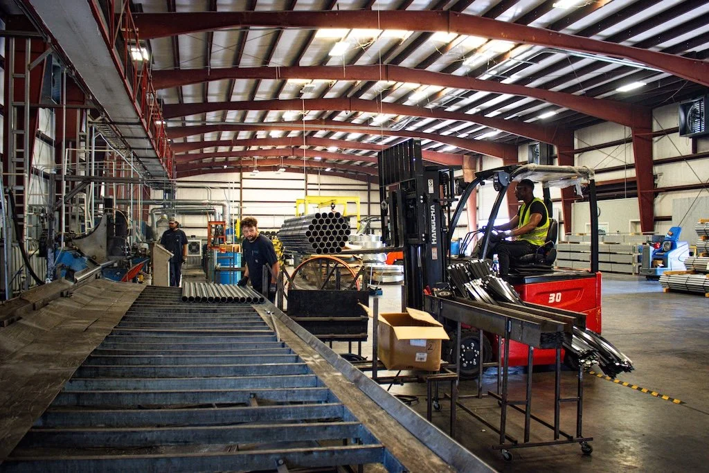 Workers in a manufacturing warehouse using forklifts and assembly lines, with metal pipes and equipment.