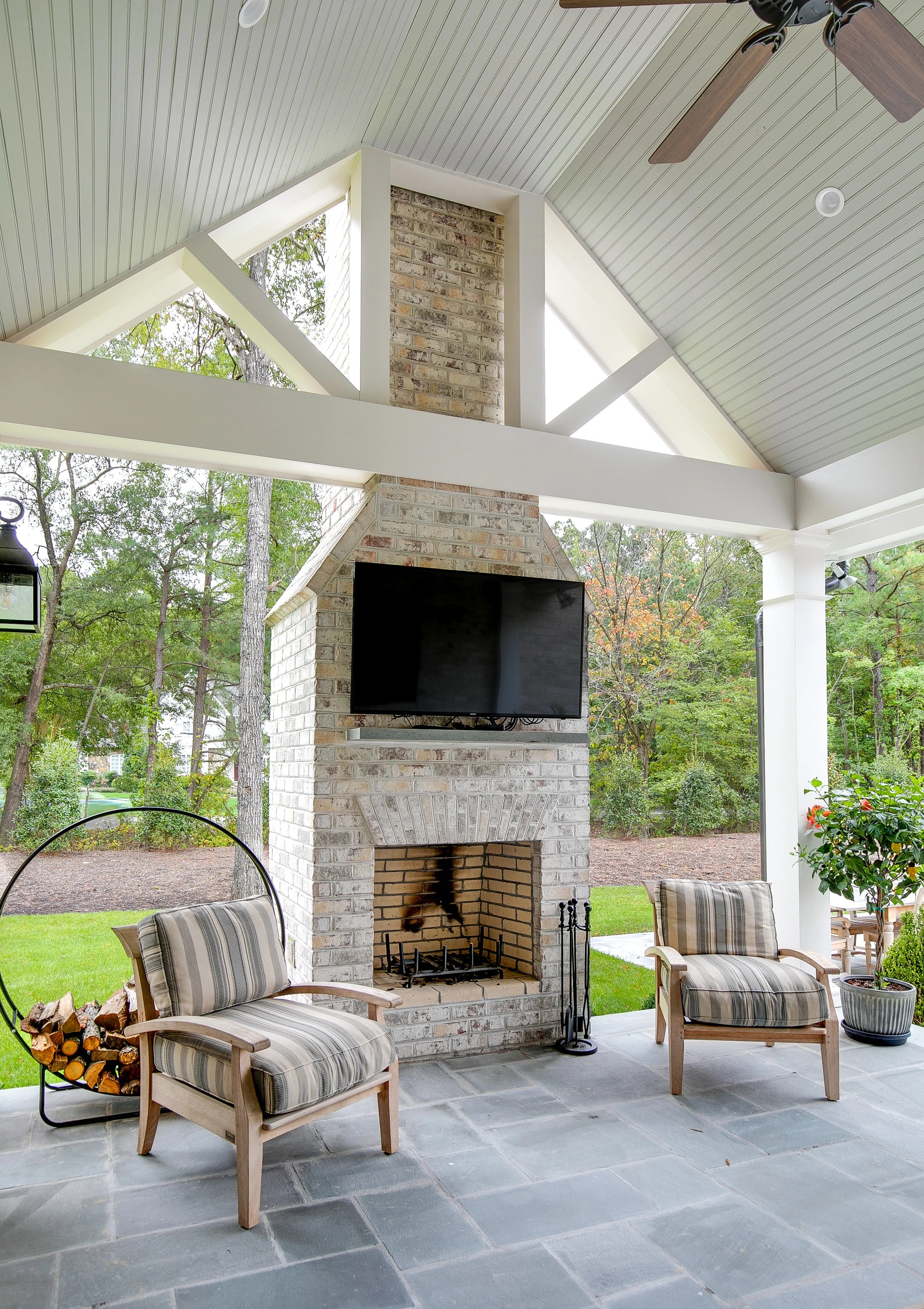 Covered patio with brick fireplace, TV mounted on the chimney, two cushioned armchairs, wood pile, potted plant, and ceiling fan, overlooking a backyard with trees.