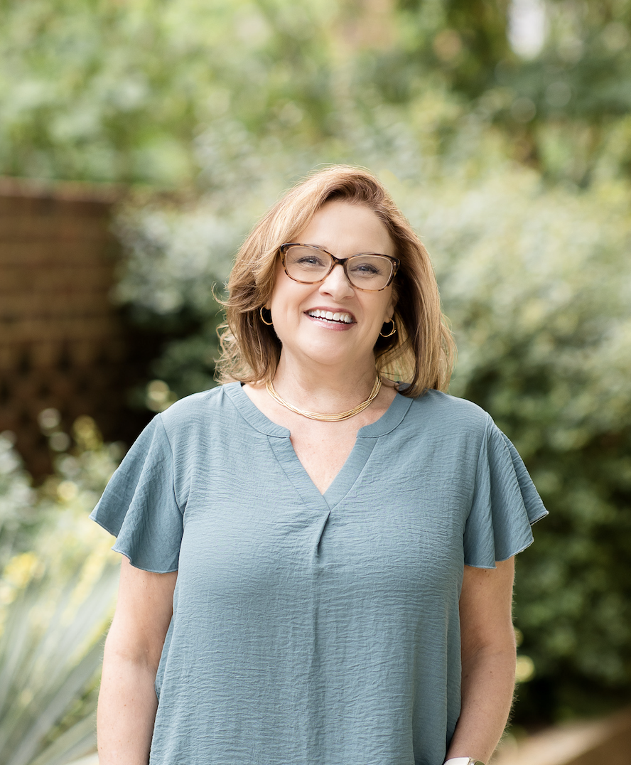 A woman with short curly hair smiling and leaning against a stone wall outdoors on a sunny day.