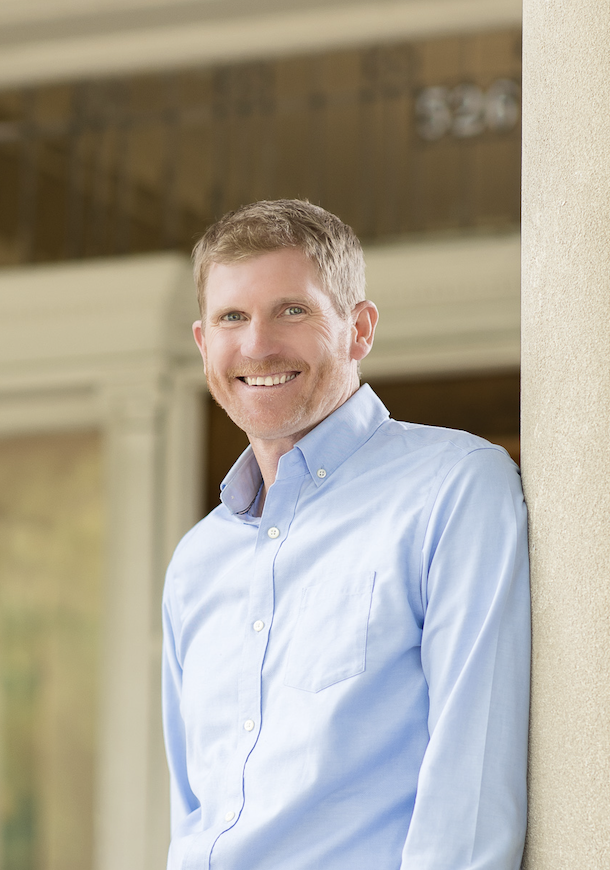 A smiling man with short brown hair and a beard, wearing a light blue button-up shirt, standing outdoors with blurred green trees in the background.