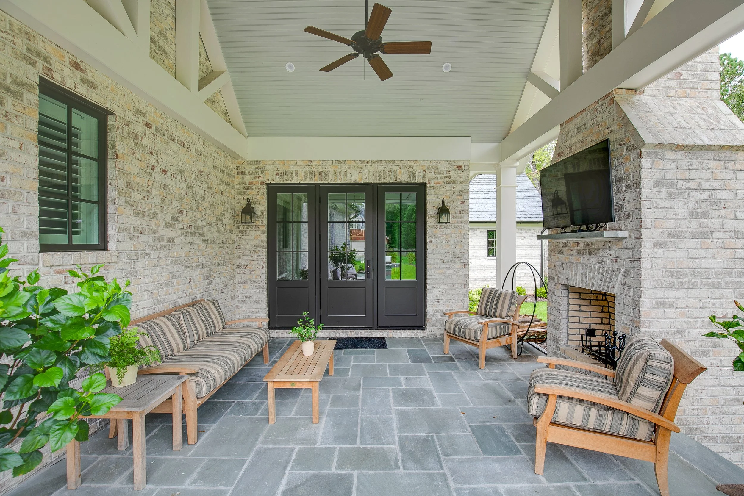Covered outdoor patio with brick walls, black double doors, and black-framed windows. Contains a striped sofa, two striped armchairs, a coffee table with a potted plant, a small wooden side table, a propane fireplace, a mounted flat-screen TV, ceilin