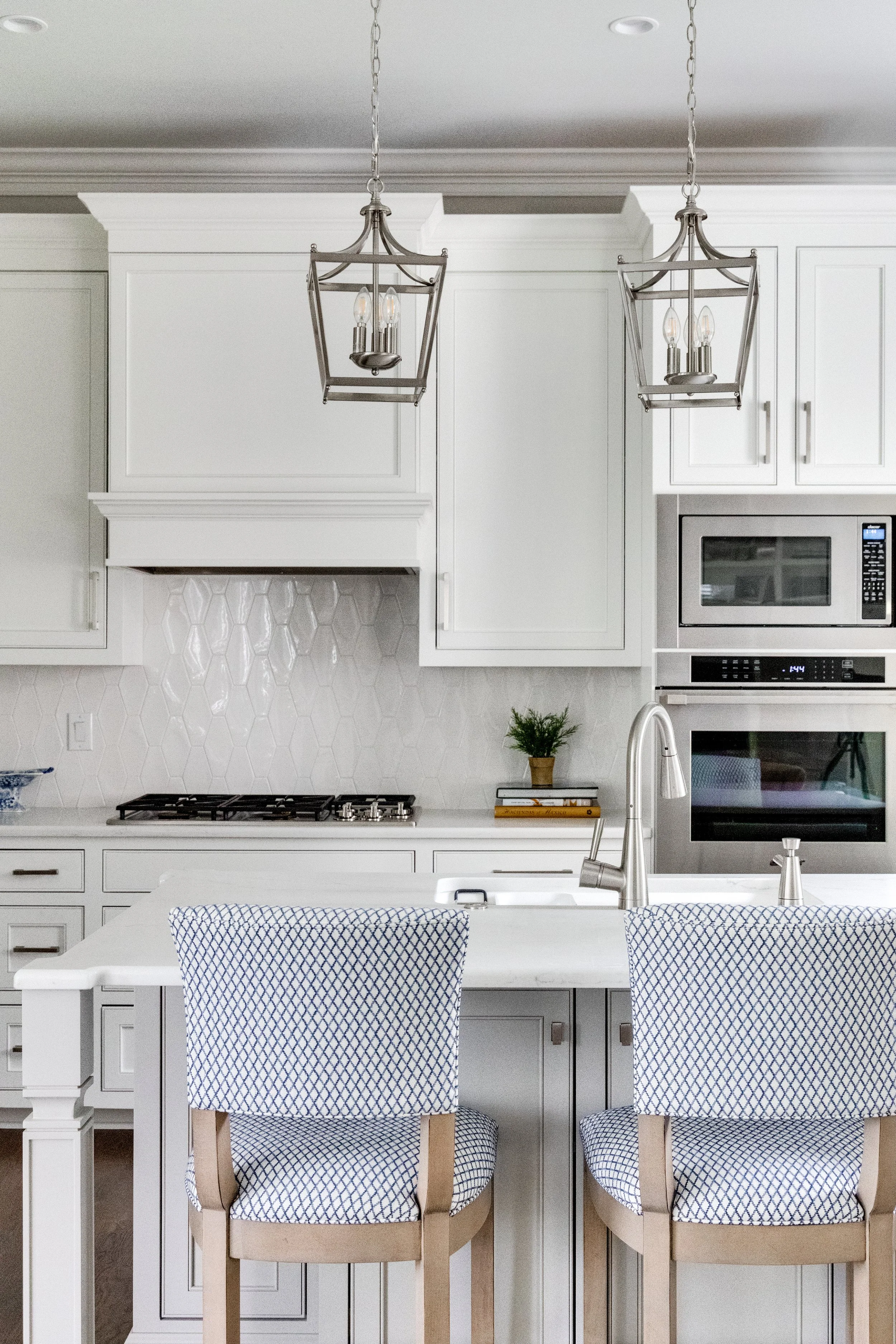 Modern white kitchen with two hanging lantern-style light fixtures, white cabinets, a built-in microwave and oven, a small potted plant, and two chairs with blue and white patterned cushions at a kitchen island.