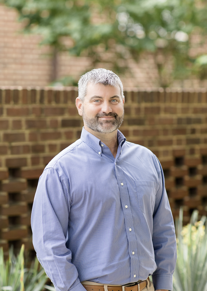 A smiling man with a beard and short dark hair, wearing a blue gingham button-down shirt and beige pants, leaning against a beige stone column outdoors.