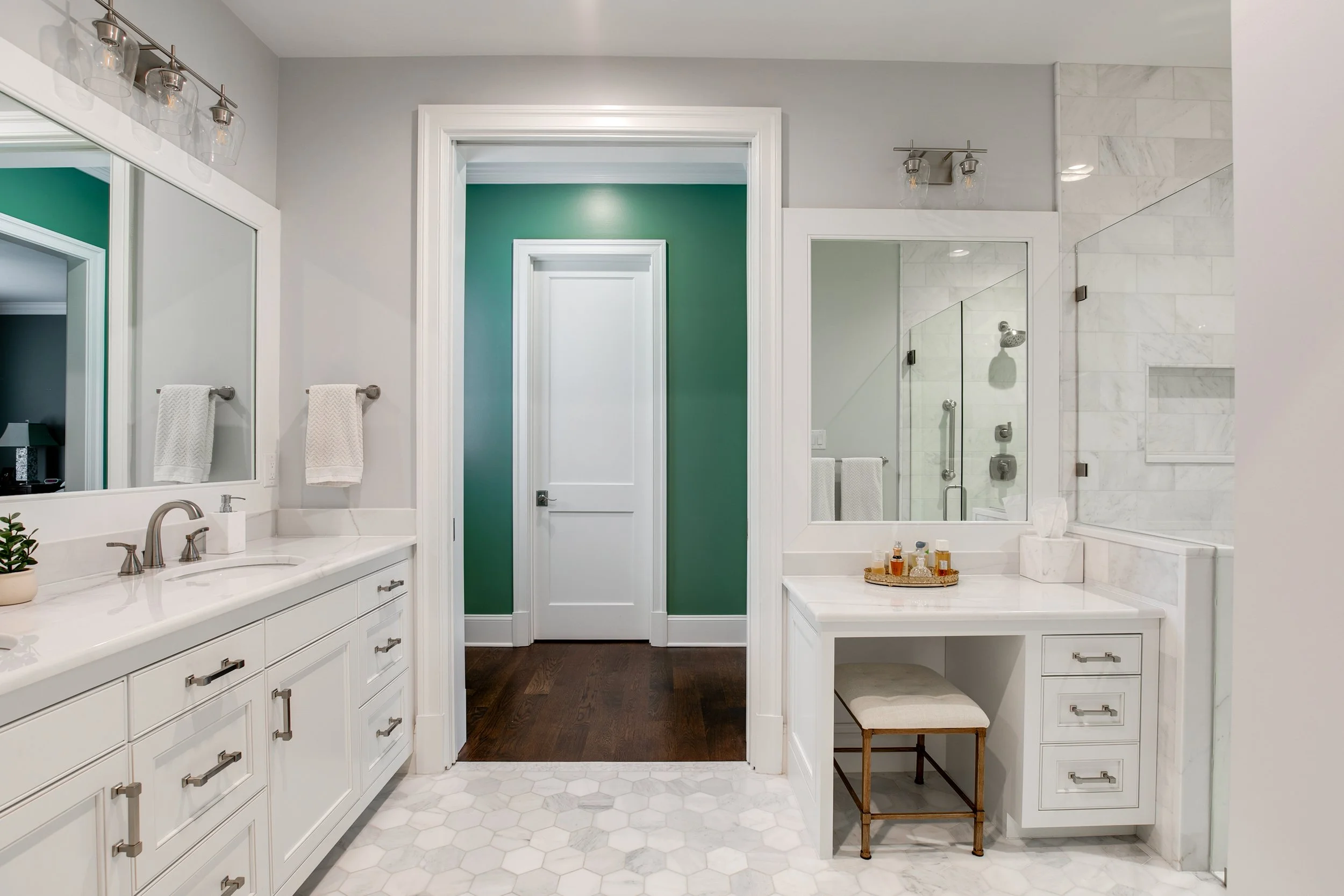 Modern bathroom with white cabinetry, marble countertops, a large mirror, and a walk-in shower with a glass door and marble walls. There are towels on towel bars and a small seating area with a stool and decor bottles.