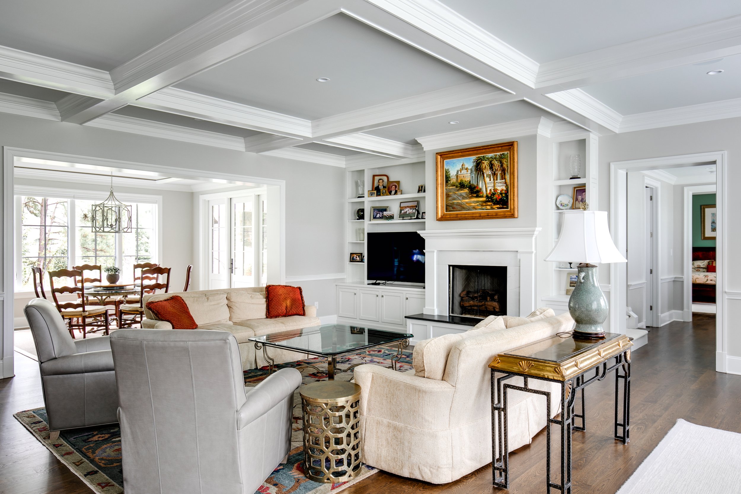 Living room with white walls, ceiling molding, two beige sofas, two white armchairs, a glass coffee table, a side table with a lamp, a rug, a fireplace, and a wall-mounted flat-screen TV. A dining area with a chandelier and wooden chairs is visible t