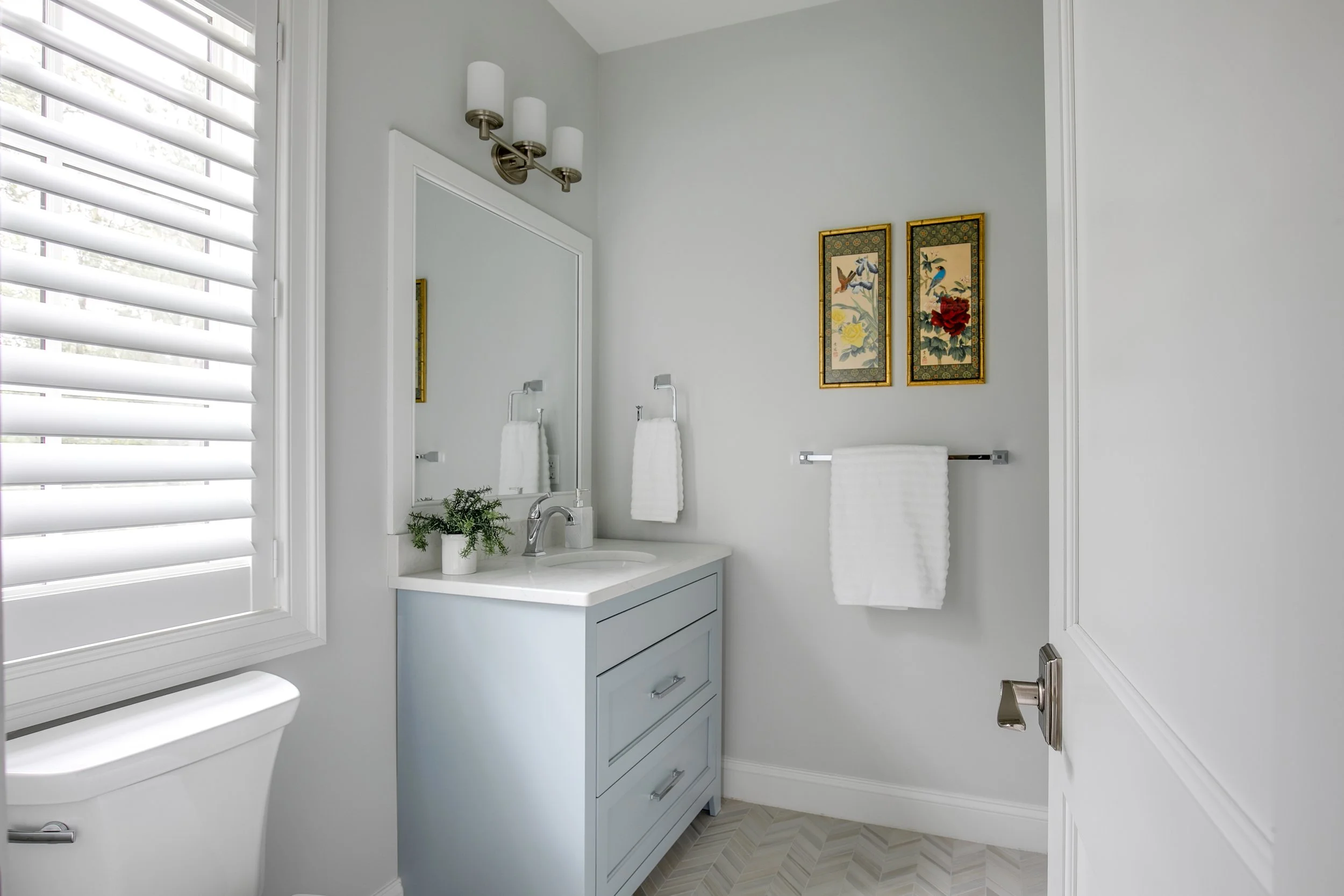 A small, modern bathroom with white walls, a blue vanity with a white countertop, a mirror, a two-light wall sconce, a small potted plant, a towel on a silver rack, two framed art pieces with flowers, a window with white shutters, and a toilet.