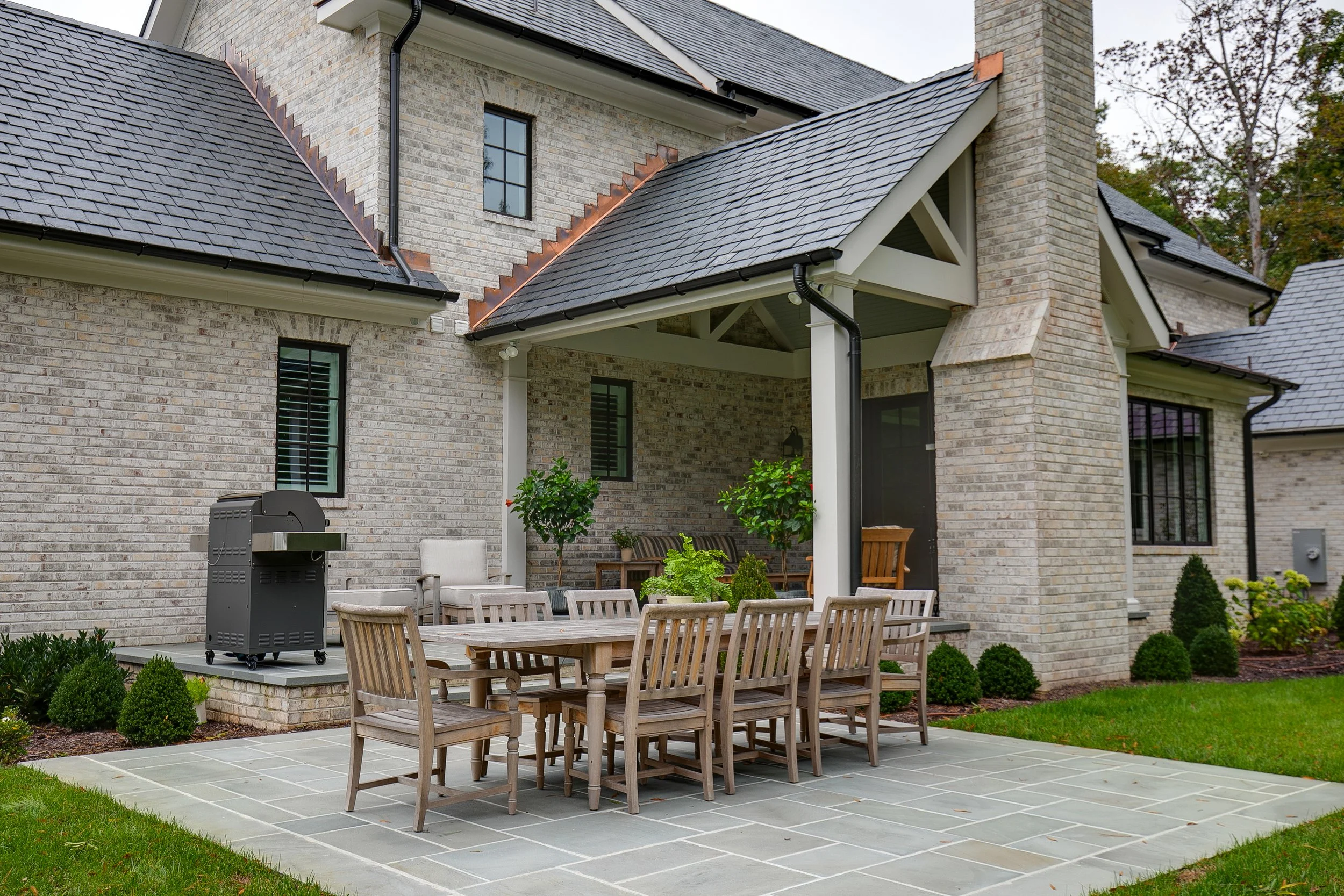 A backyard patio with a long wooden table and multiple chairs, a grill, potted plants, and a brick house with a covered porch and multiple windows.