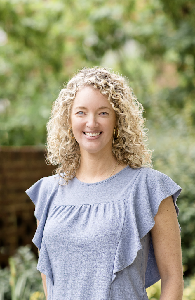 A woman with curly blonde hair smiling outdoors, wearing a light blue ruffled dress and gold jewelry.