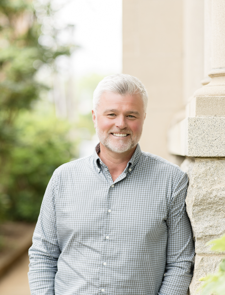 Portrait of a smiling middle-aged man with gray hair and beard, wearing a light blue checkered shirt, standing outdoors with greenery and a brick building in the background.