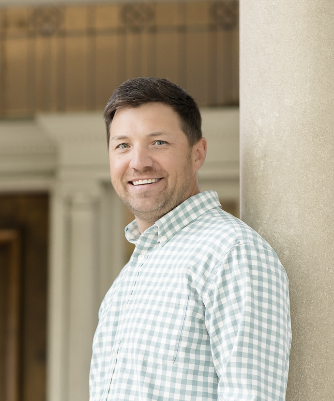Smiling man in a blue checkered button-up shirt outdoors with blurred green trees in the background.