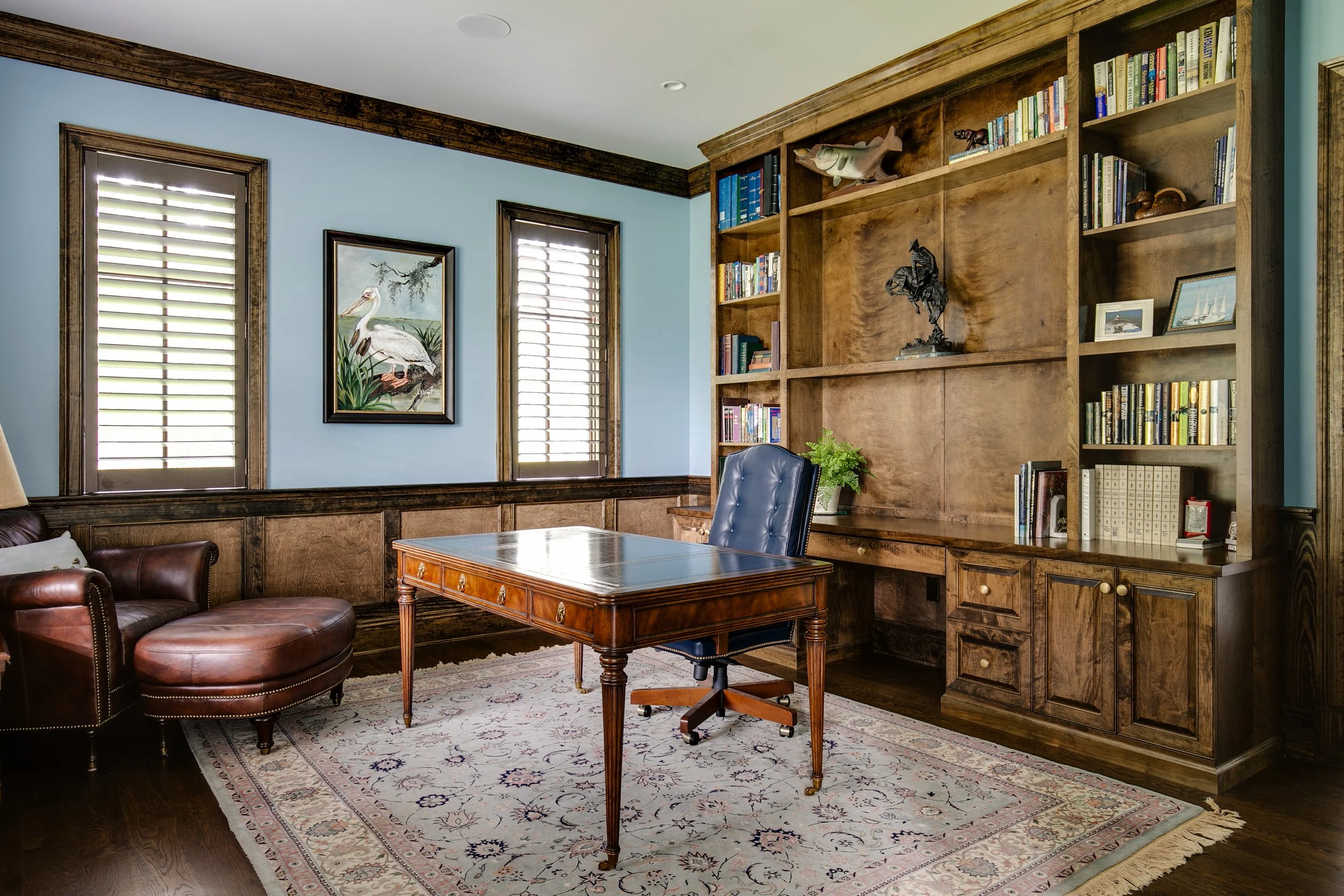 Home office with wooden furniture, a leather armchair, a painting of a heron, and a large bookshelf filled with books, decorative items, and framed photos.