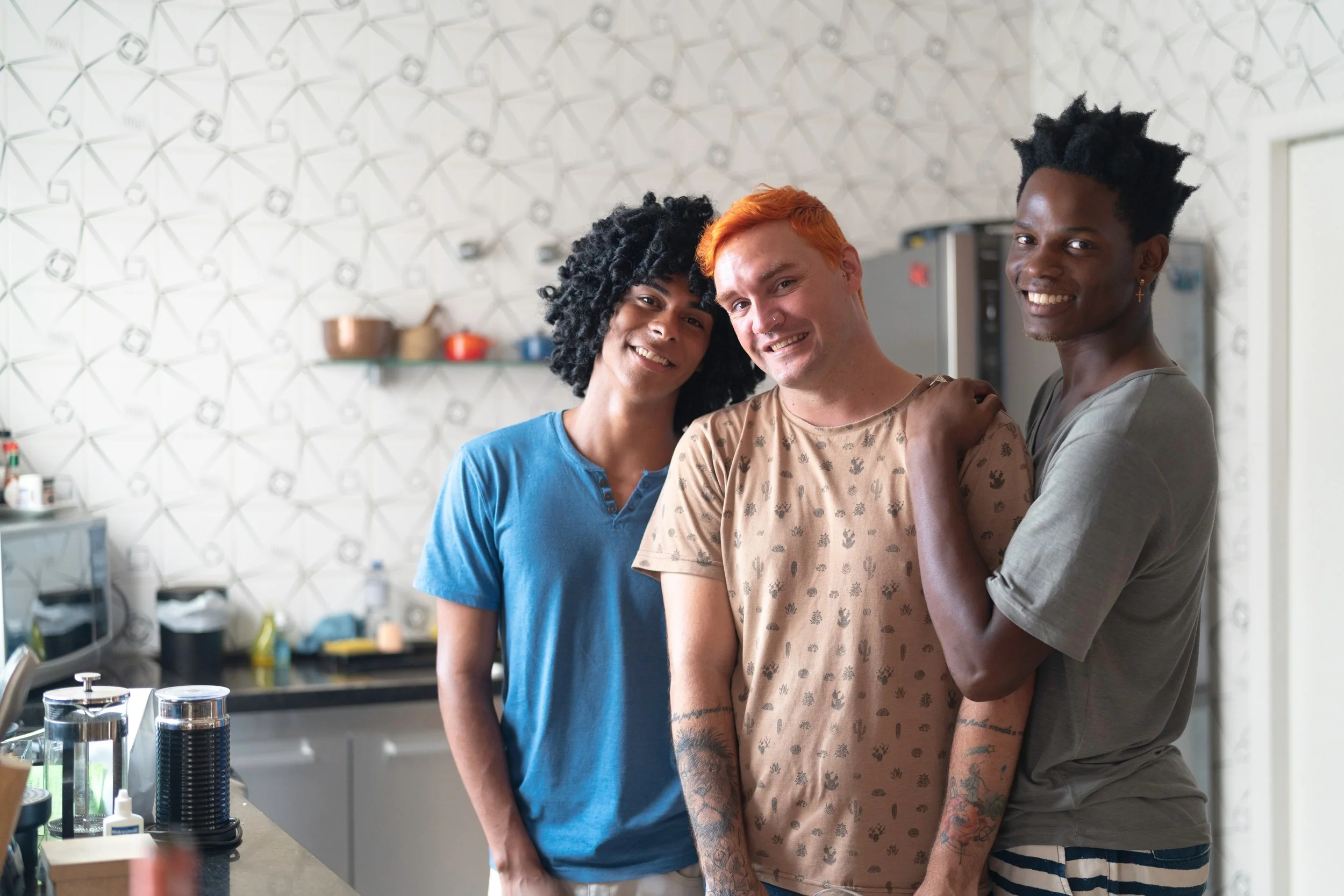Three young adults smiling together in a kitchen, standing close with arms around each other, in a modern, patterned background setting.