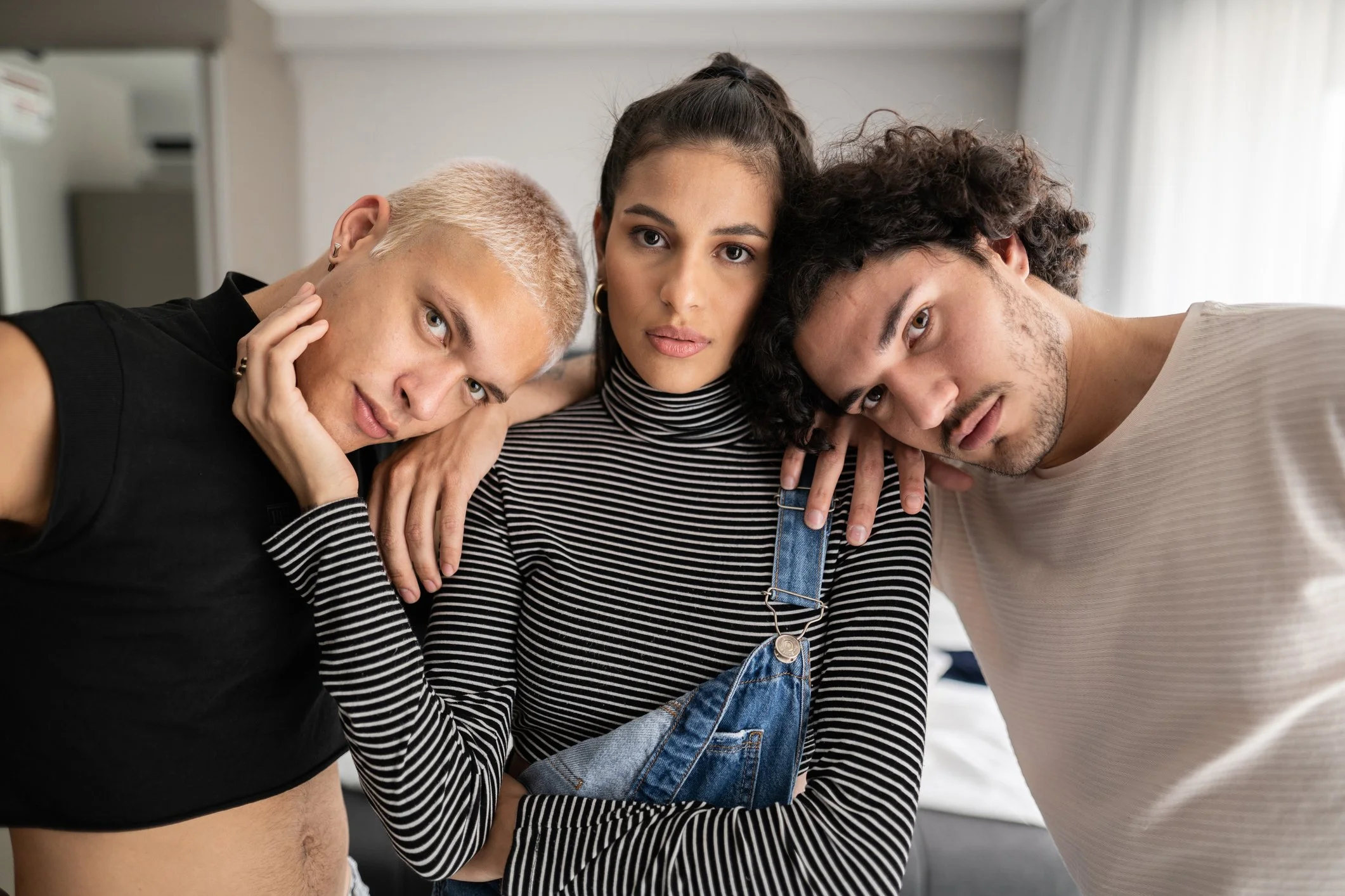 Three young adults posing close together indoors, with their heads leaning in and hands touching each other's shoulders or face, looking at the camera.