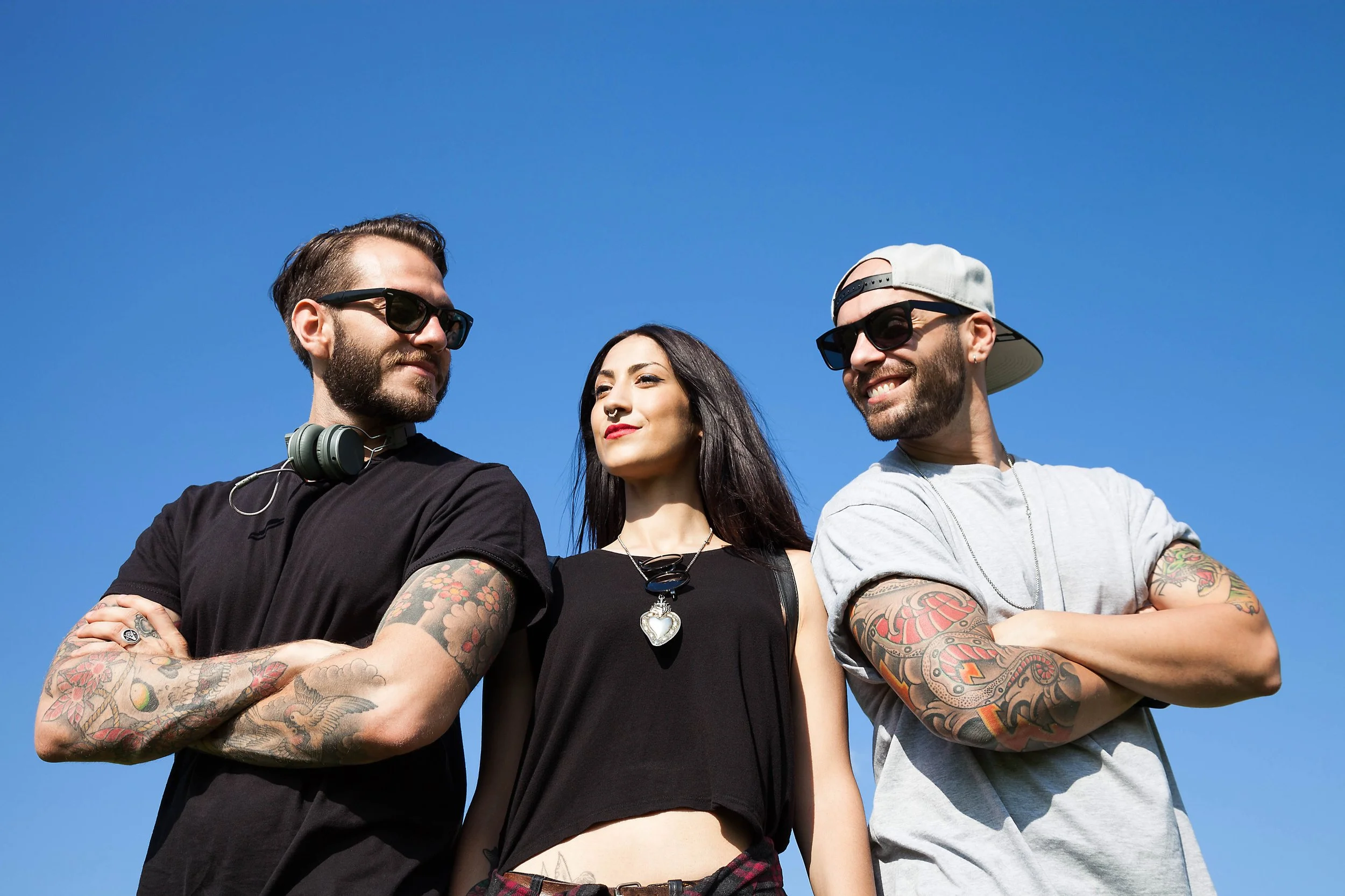 Three young adults standing outside under a clear blue sky, with two men wearing sunglasses and tattoos, and a woman with long dark hair in the middle.