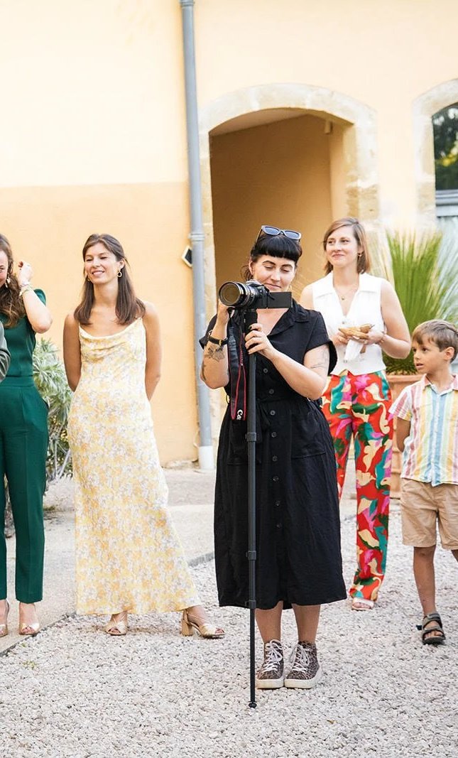 A woman in black dress and sneakers takes a photo with a professional camera on a tripod, surrounded by three women and a boy outdoors near a yellow building.