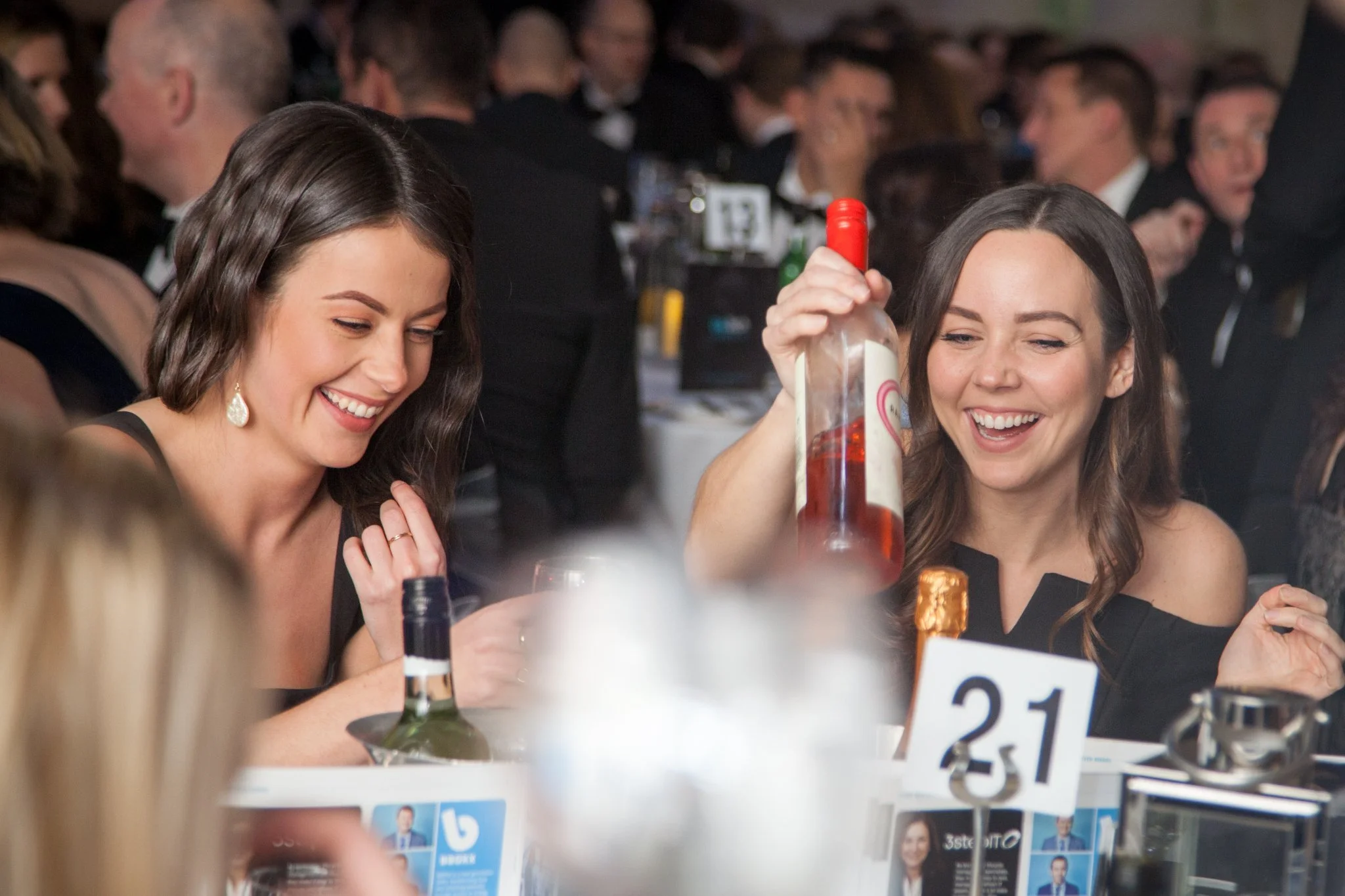 Two women smiling and laughing at a table during a formal event, one holding a bottle of rosé wine.