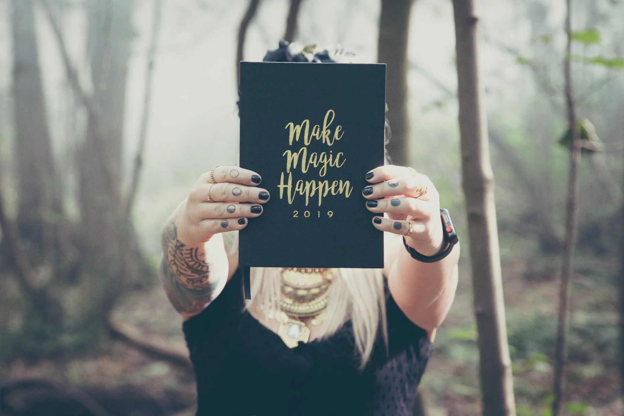 Person holding a black notebook with gold text saying 'Make Magic Happen 2019' in a forest setting.