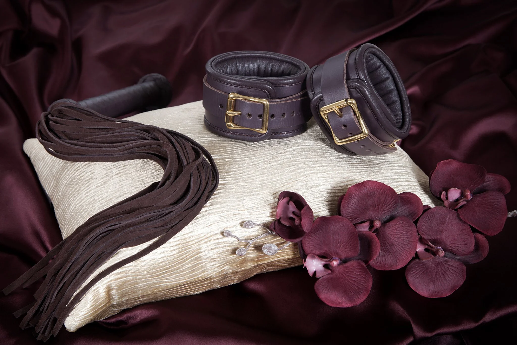 A BDSM-themed display with a wooden paddle, adjustable leather cuffs with gold buckles, a leather riding crop, a pillow with a textured cream cover, a cluster of burgundy orchids, and a small silver jewelry piece with clear stones, set on a dark sati