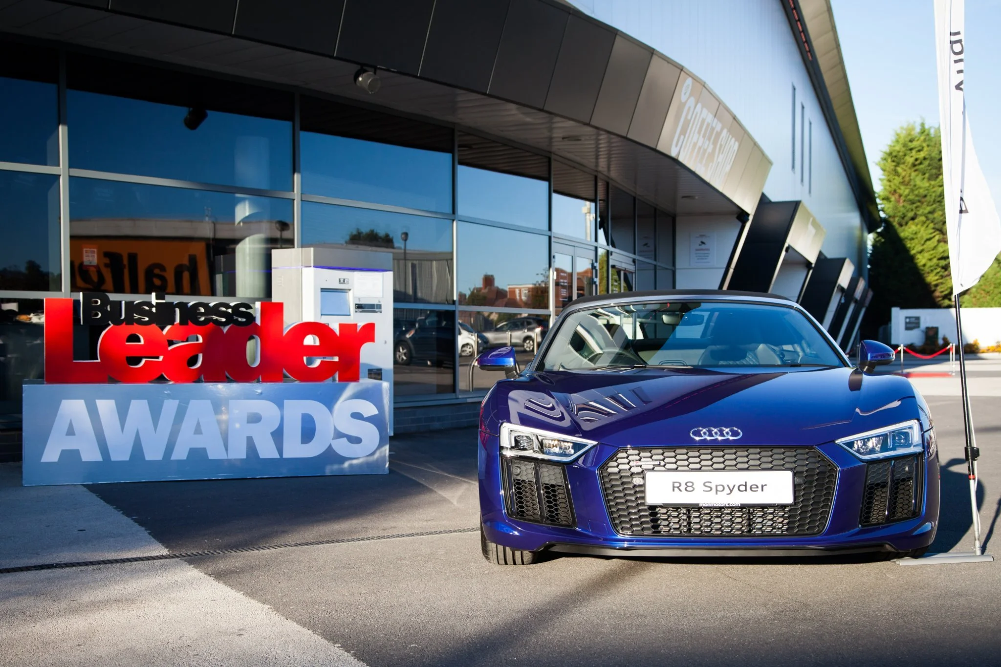 Blue Audi R8 Spyder convertible parked outside a building with large glass windows. A sign that reads 'Business Leader Awards' is near the car, with a flag on the right side.