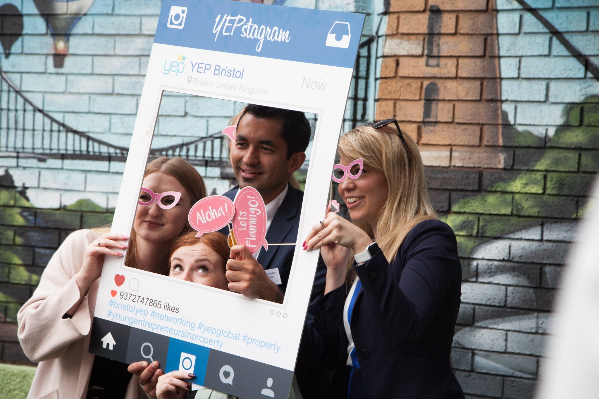 Group of four people holding up a large social media frame prop at a party, wearing fun pink and white glasses and holding signs that say "Aloha!" and "Let's flamin go!" against a graffiti wall background.