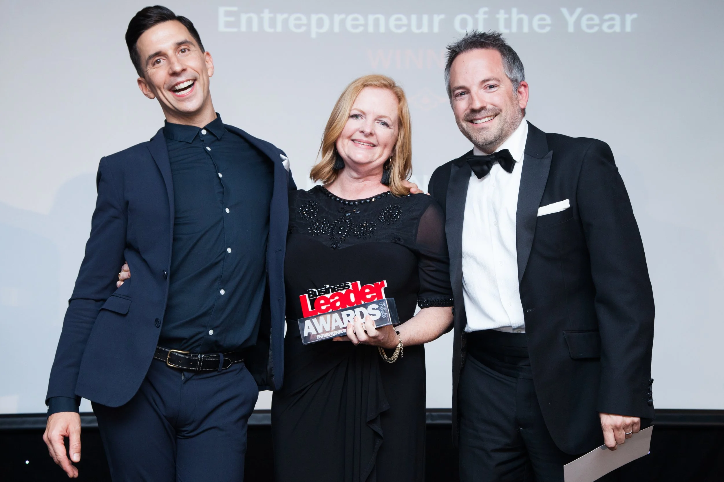 Three people dressed in formal attire posing together on stage at an award ceremony, with a woman holding a plaque or award that says "Business Leader Awards." The background features a screen with text that says "Entrepreneur of the Year."
