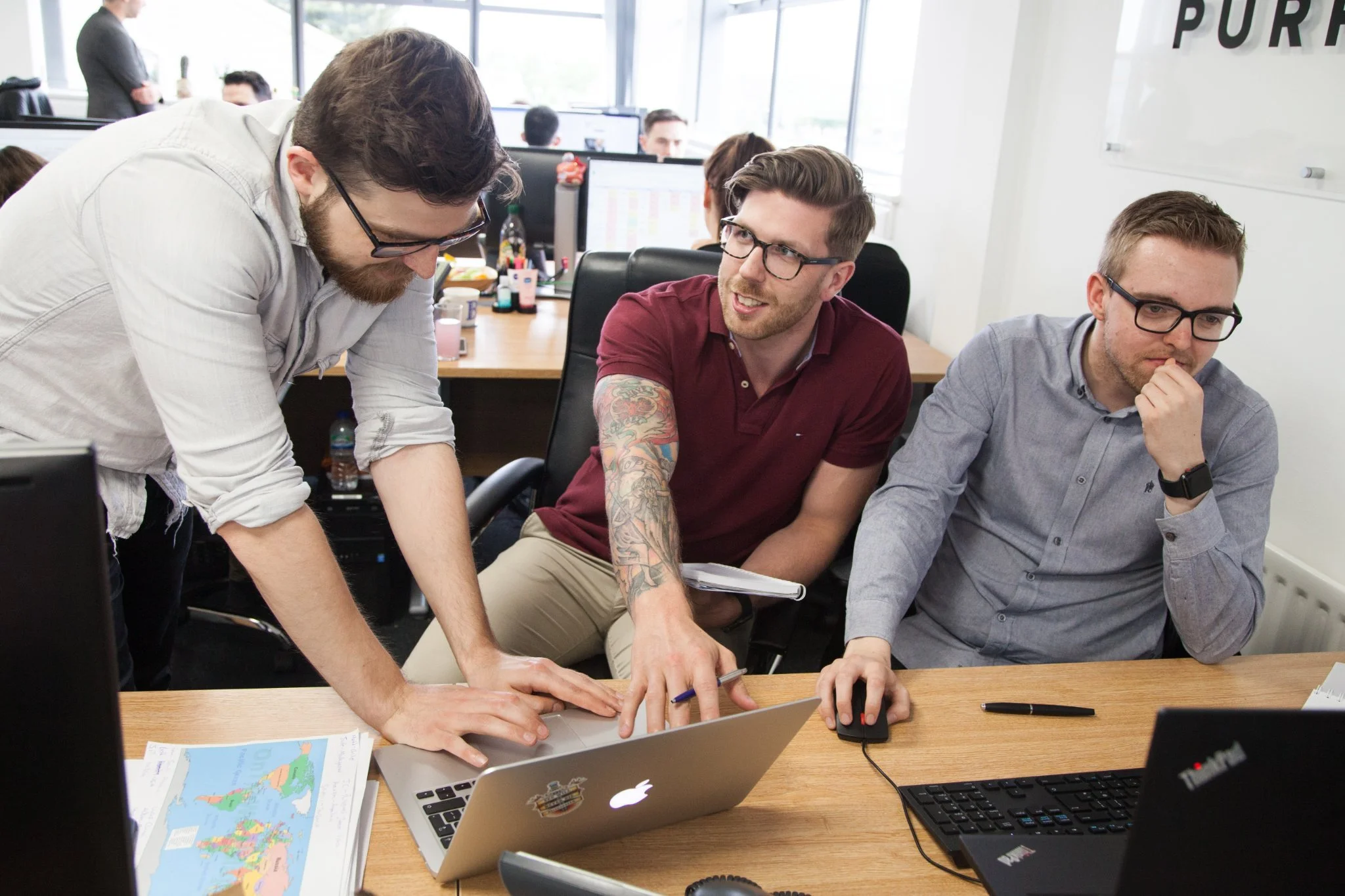 Three men working on a laptop in an office, two sitting and one standing, with multiple computer screens and office supplies around.