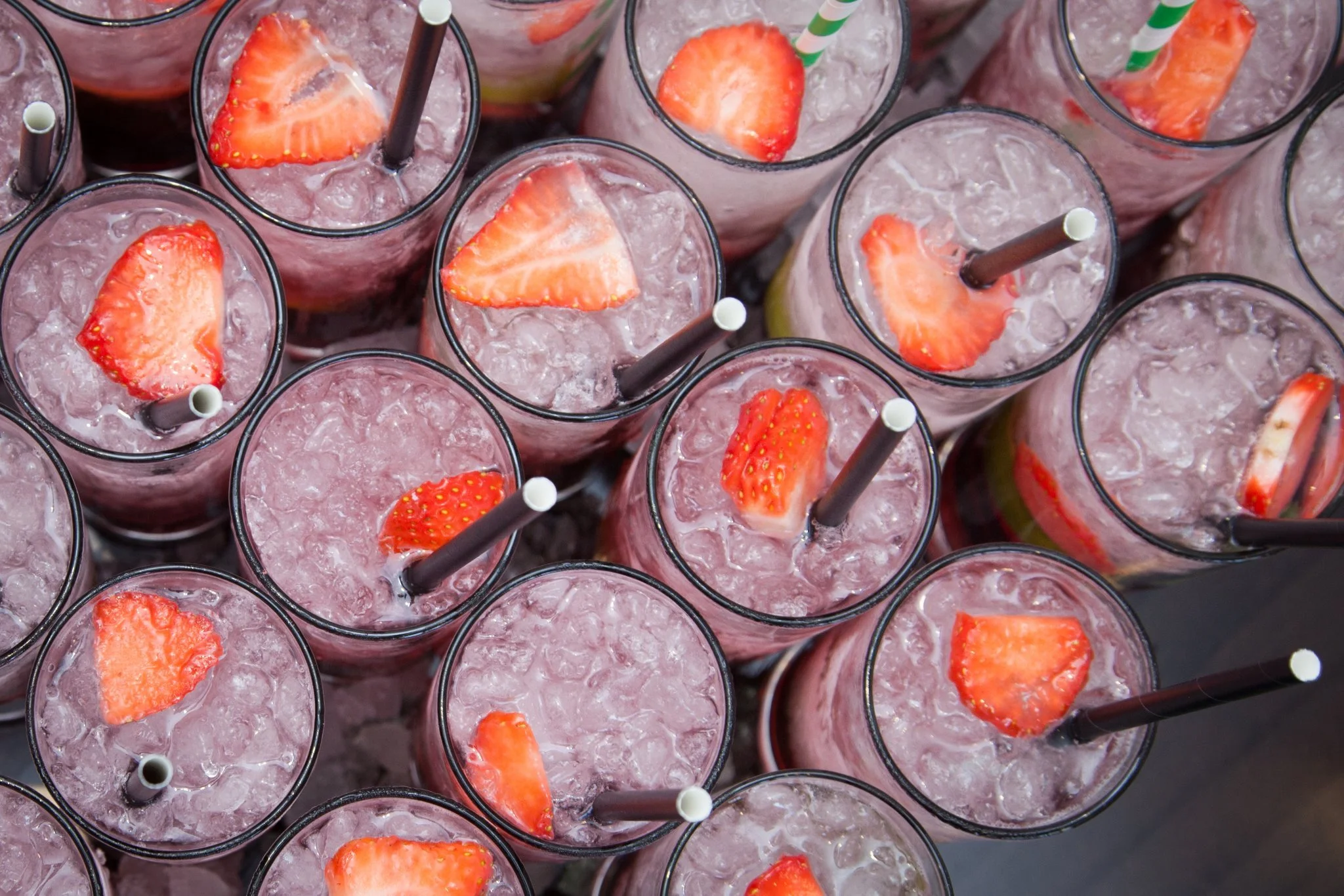 Multiple glasses filled with pink strawberry drinks with ice, with strawberry slices and brown straws on top.