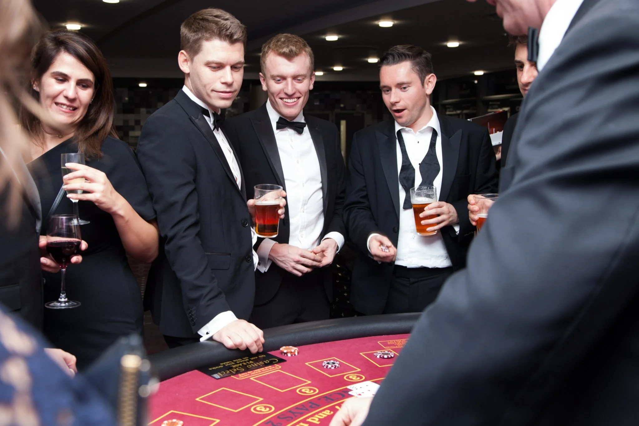 Group of people in formal attire playing at a casino table with drinks in hand.
