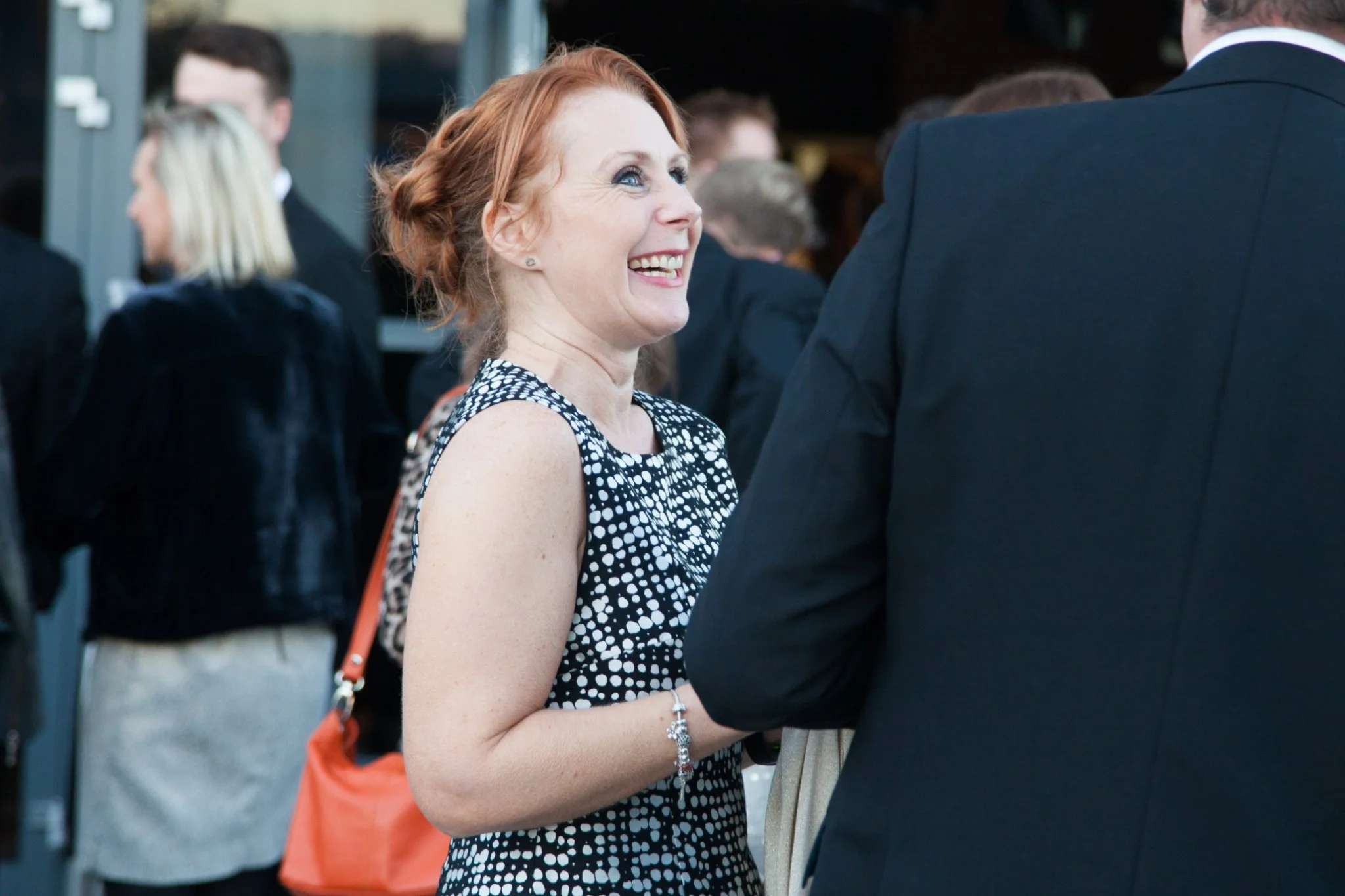 A woman with red hair smiling and engaging in conversation at an event, wearing a sleeveless black and white polka dot dress, with a background of people in formal attire.