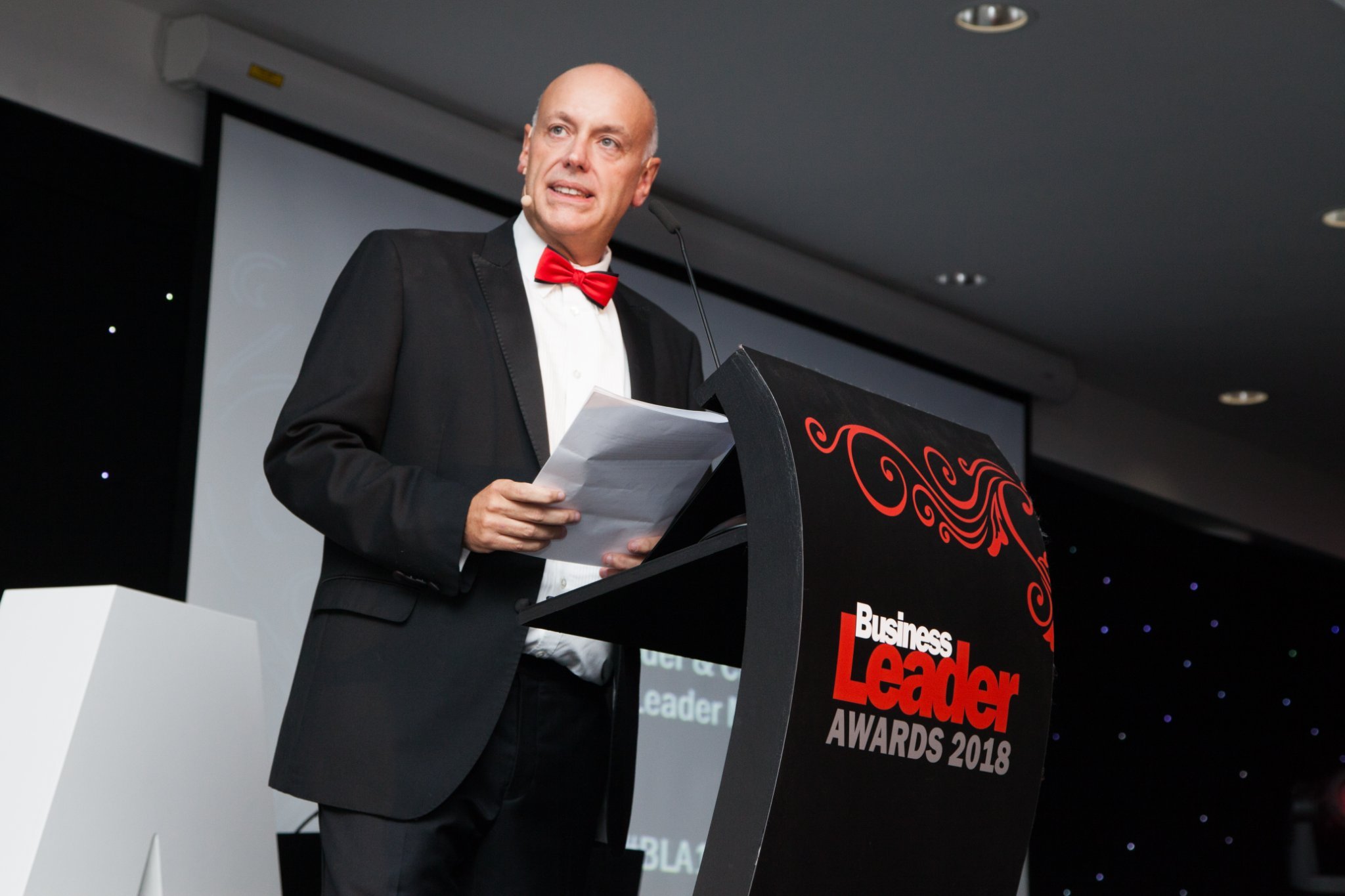 A man in a tuxedo with a red bow tie speaking at a podium during the Business Leader Awards 2018.