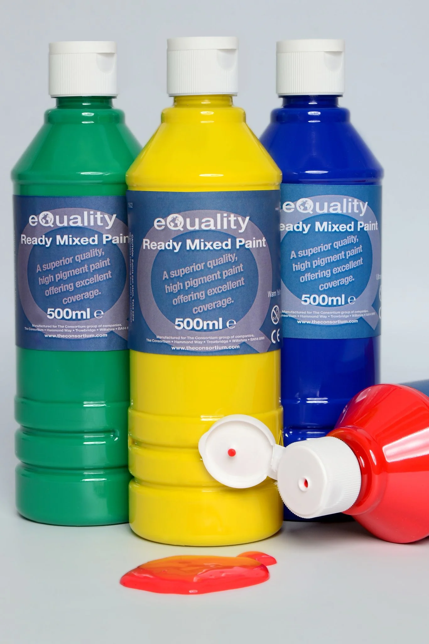 Three bottles of ready mixed paint in green, yellow, and blue, with a red bottle tipped over, spilling red paint onto a white surface.