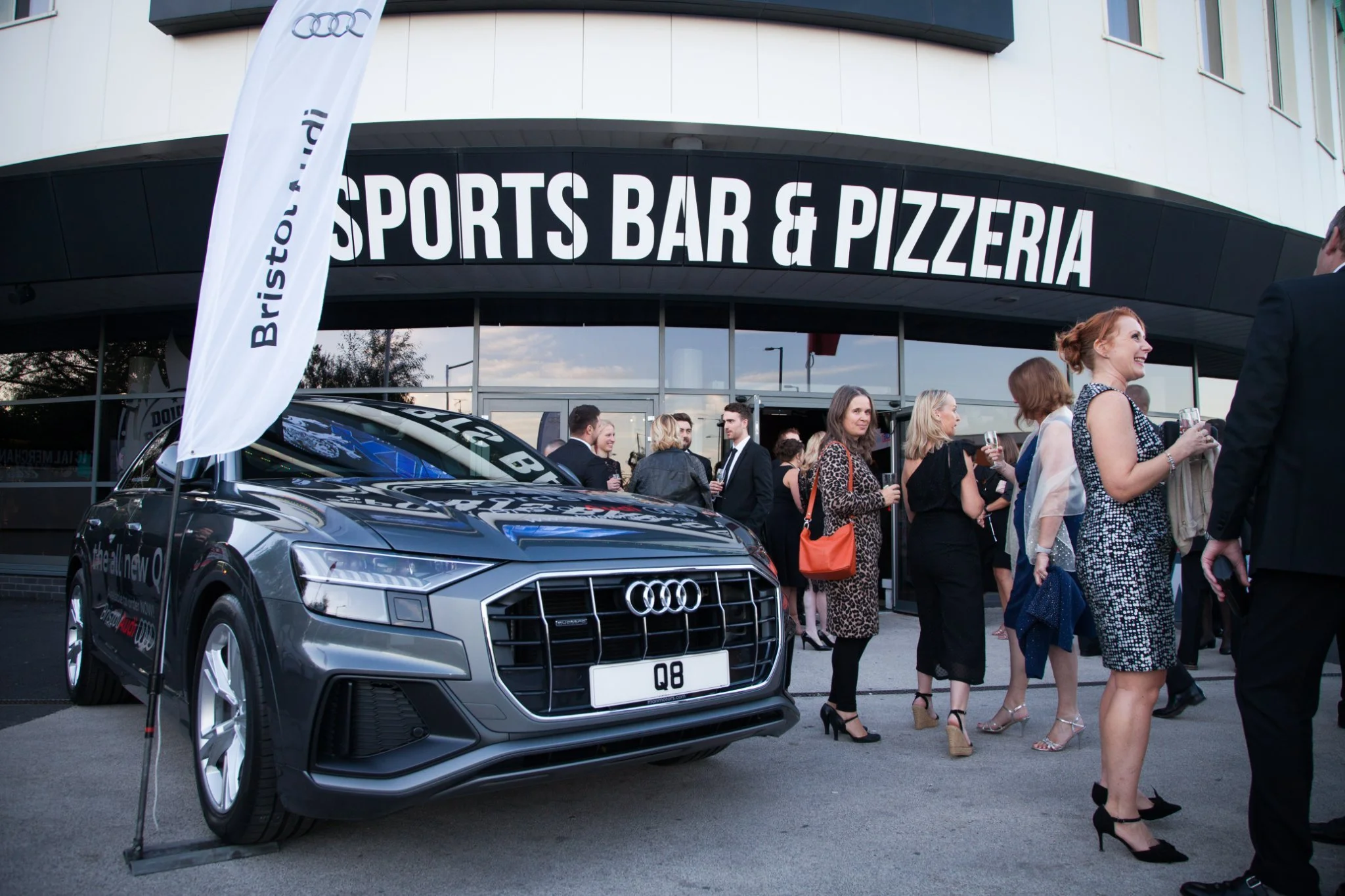 Group of people dressed in formal attire standing outside a sports bar and pizzeria, with a silver Audi Q8 SUV parked nearby and a flag with the text "Bristol" on it.
