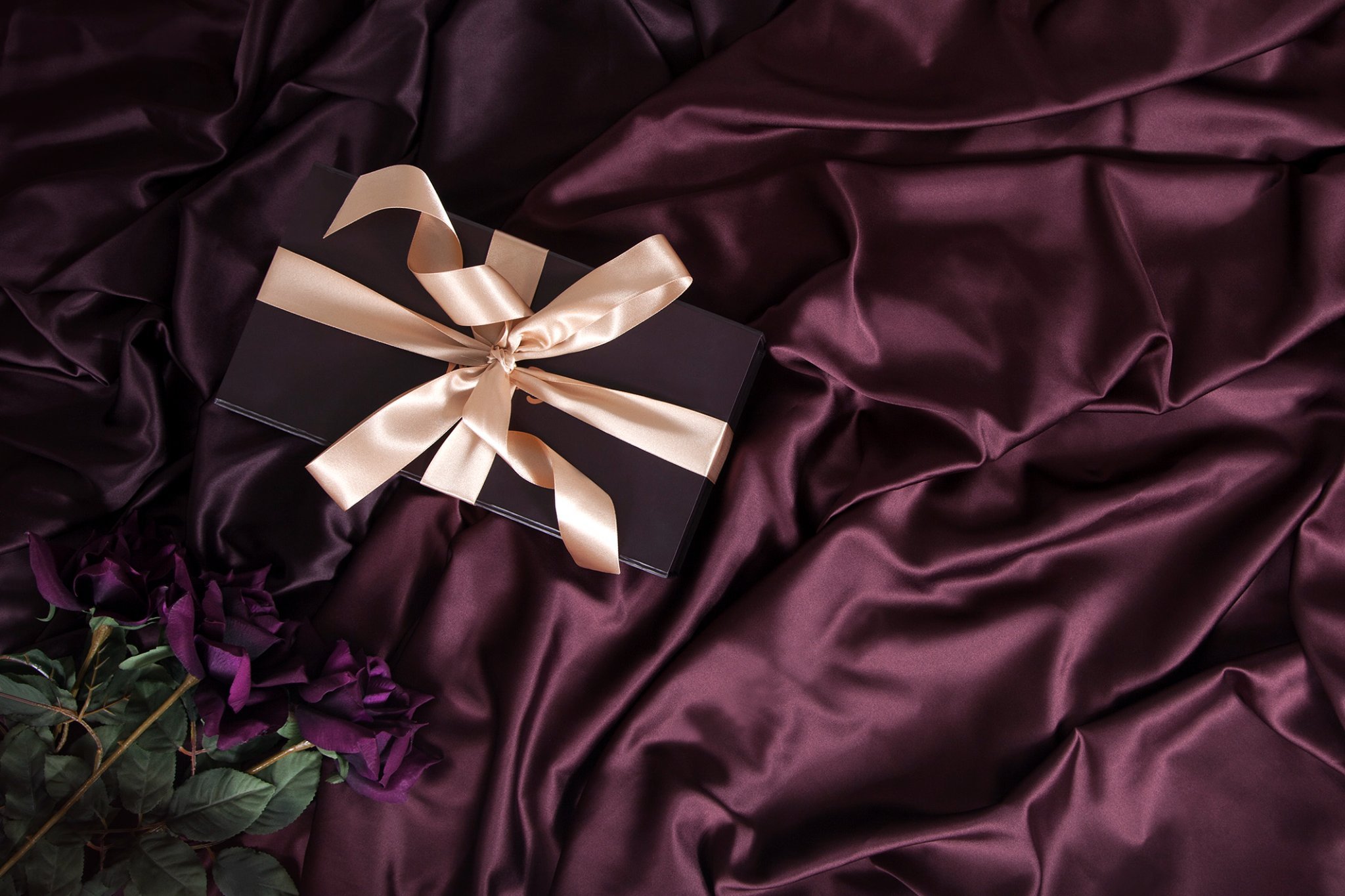 A black gift box with a beige satin ribbon bow, resting on dark purple satin fabric, with a few purple flowers nearby.