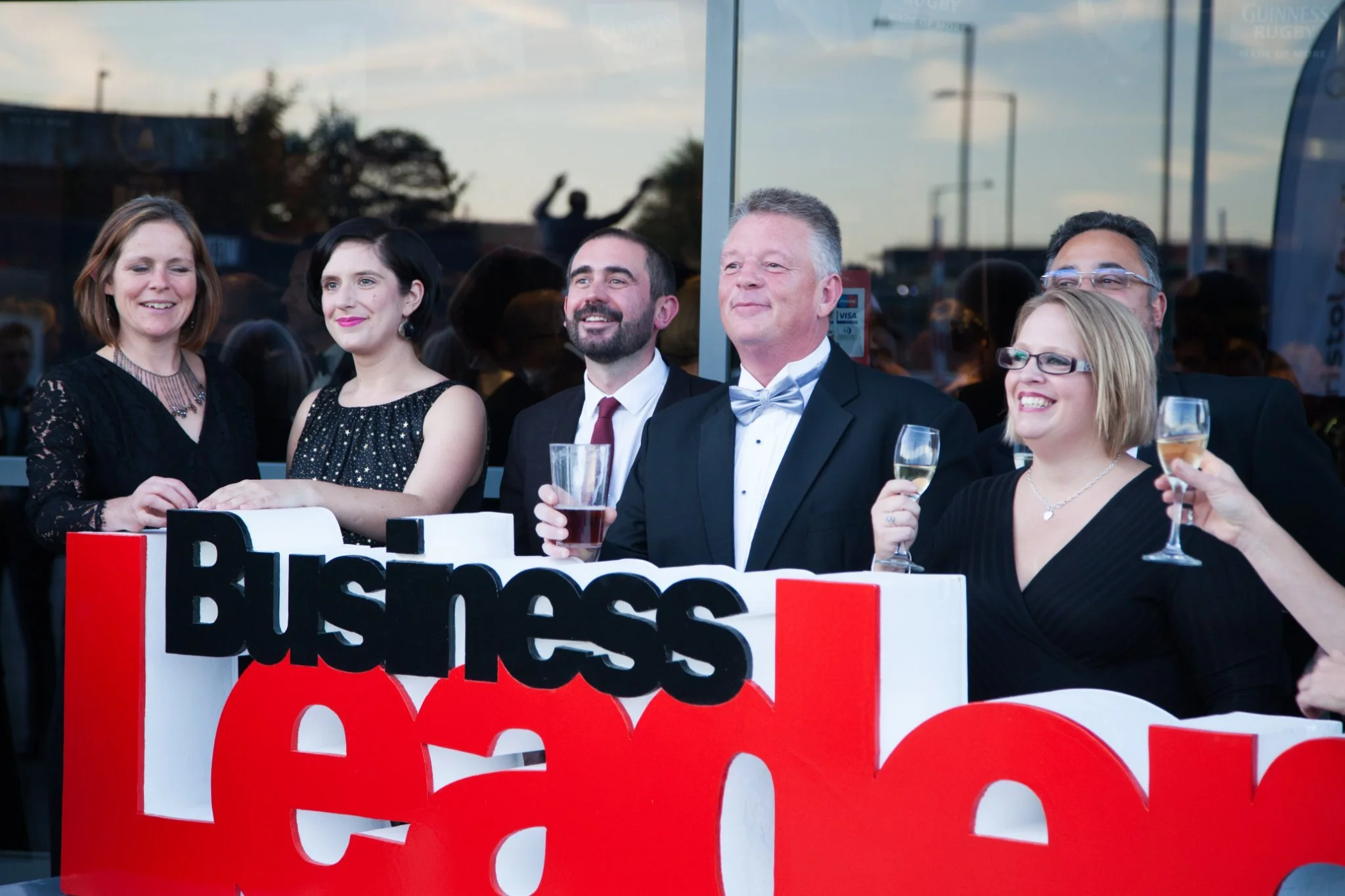 Celebrating business professionals at an outdoor event with a large sign that reads 'Business Real'. The group is dressed in formal attire and holding wine and drink glasses, smiling and enjoying the occasion.