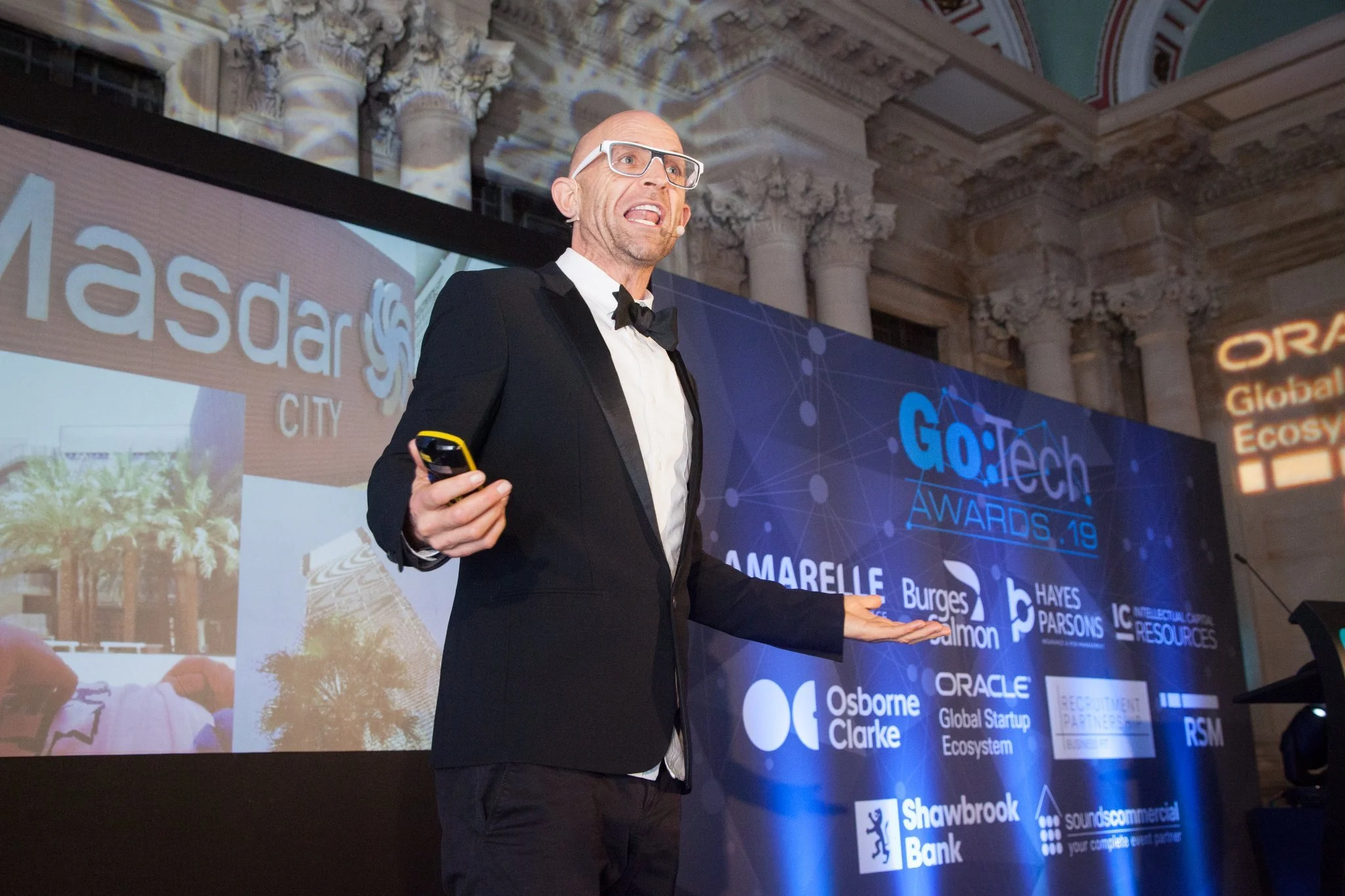 A man in a tuxedo with glasses giving a presentation on stage at the GO:Tech Awards 2019, with various sponsor logos displayed on a large screen behind him.