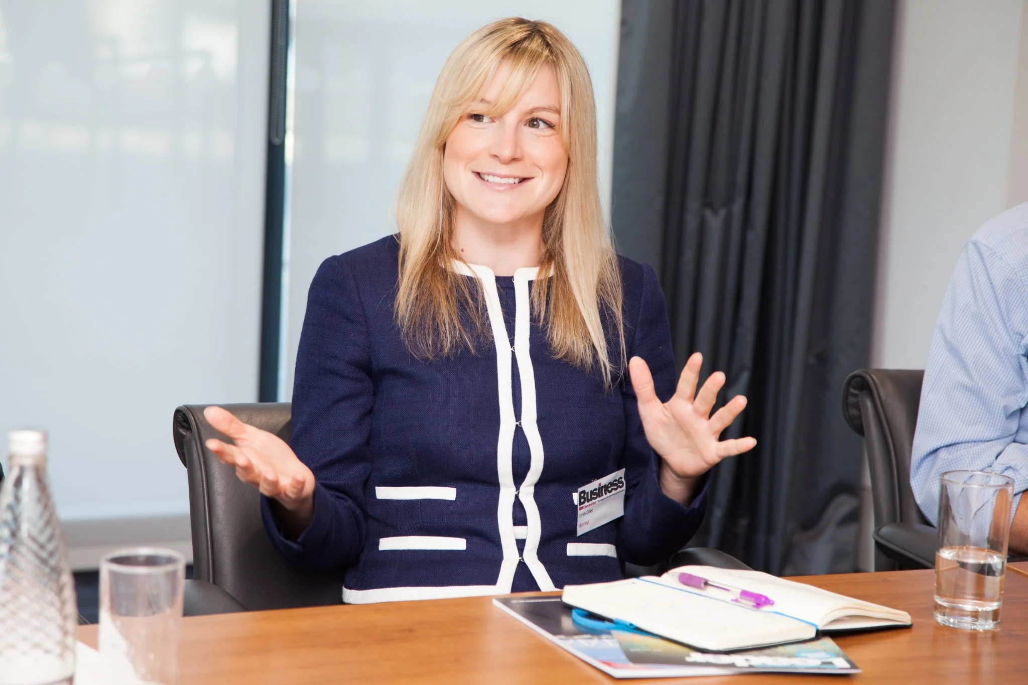 A woman with blonde hair, wearing a navy blue jacket with white trim, sitting at a conference table, smiling and gesturing with her hands during a meeting.