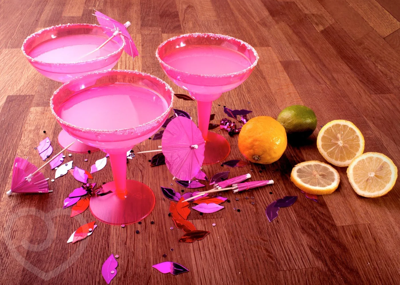 Three pink cocktails in sugar-rimmed glasses on a wooden table, decorated with pink and purple paper umbrellas and confetti, along with fresh lemons, a lime, and a lemon half.