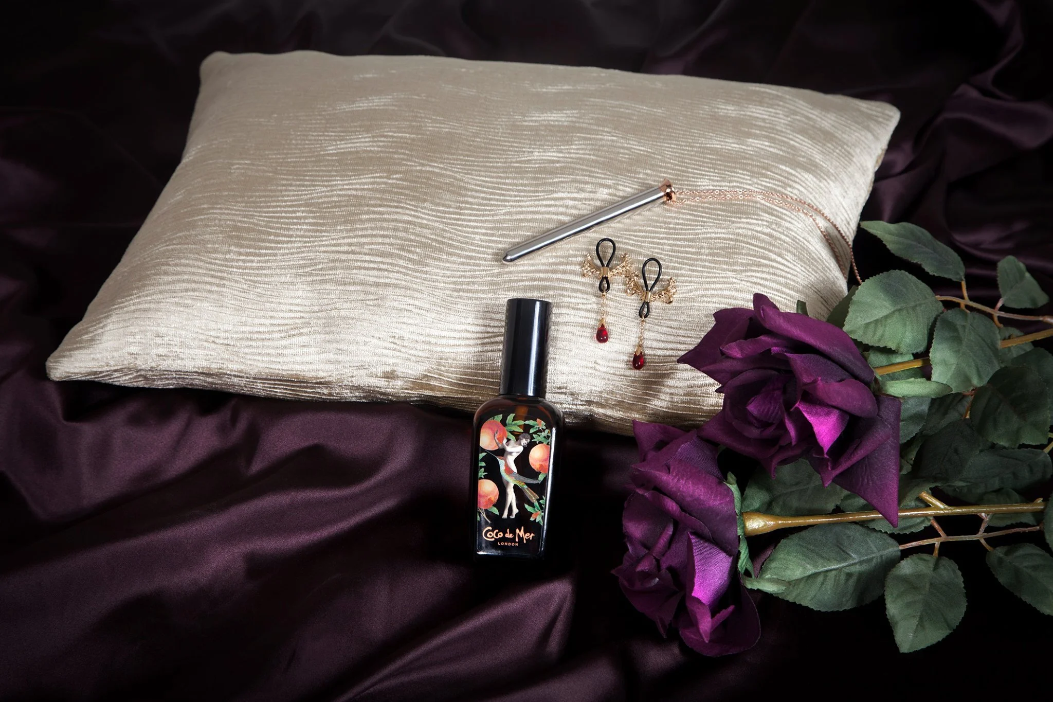 A silver pillow with a textured surface, lying on dark purple satin fabric. On the pillow are a silver pen with a chain, a pair of gold and black earrings with red gemstones, and a small dark bottle of Coco de Mer perfume with an illustrated label. T