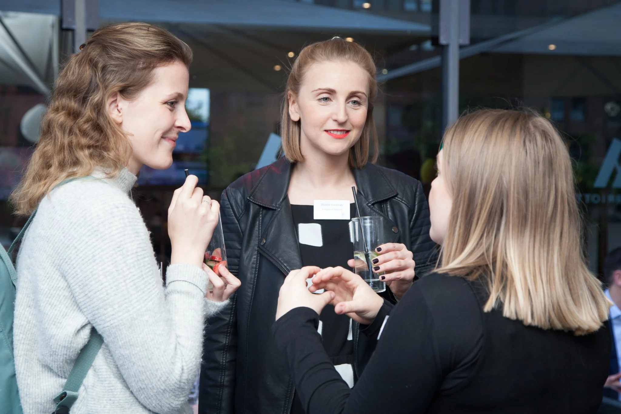 Three women engaged in conversation at a social gathering indoors during the daytime. One woman is holding a drink with lime, another woman has a drink with a straw, and the third woman appears to be speaking or making a point.