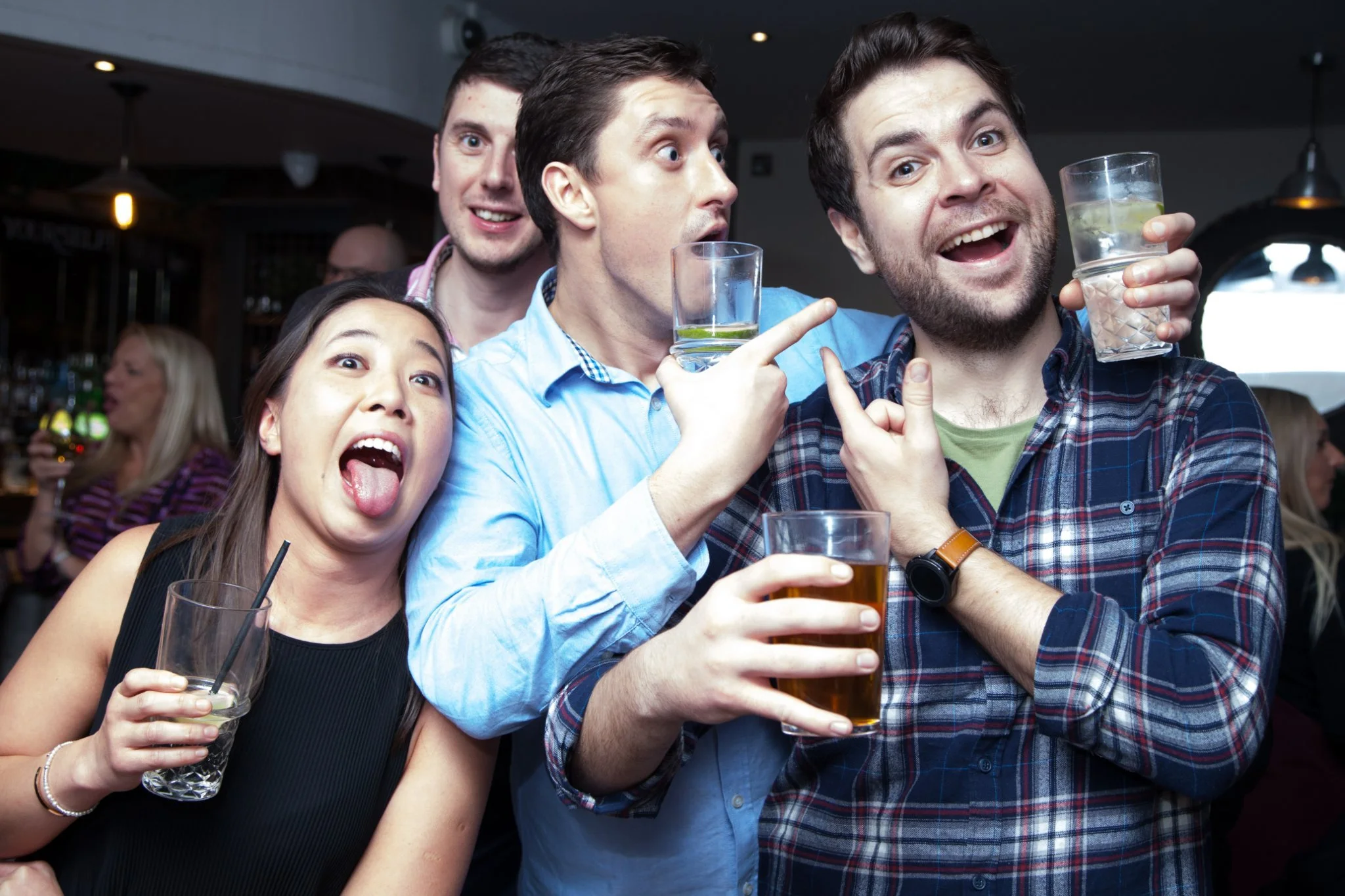 Group of four friends at a bar celebrating, holding drinks and making playful gestures, with other patrons in the background.