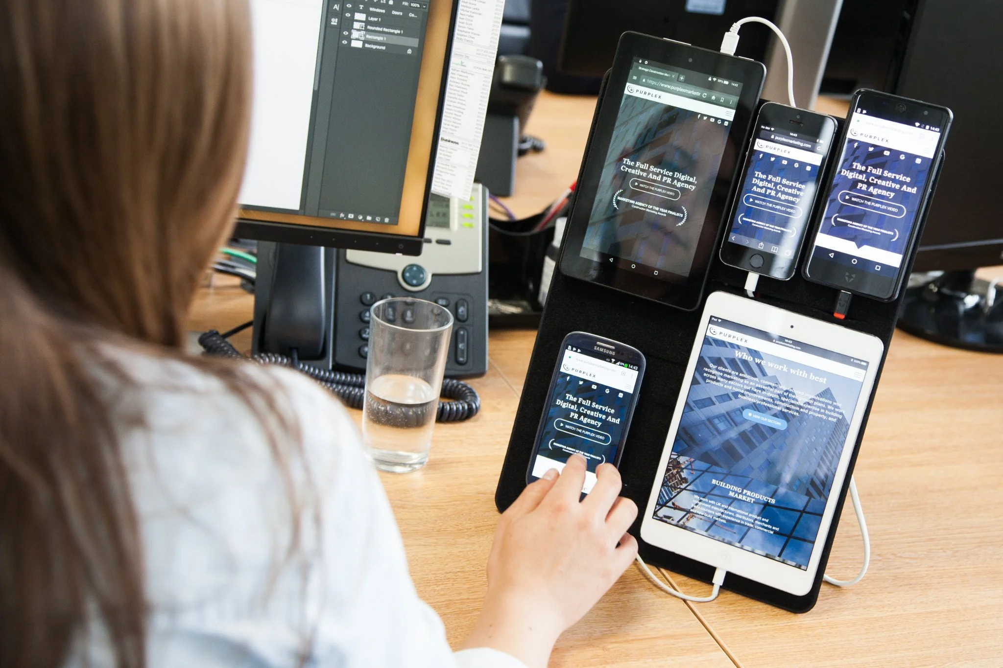 A woman working at her desk monitors multiple electronic devices including a desktop computer, a tablet, and five smartphones, all displaying the same website for a digital and PR agency.