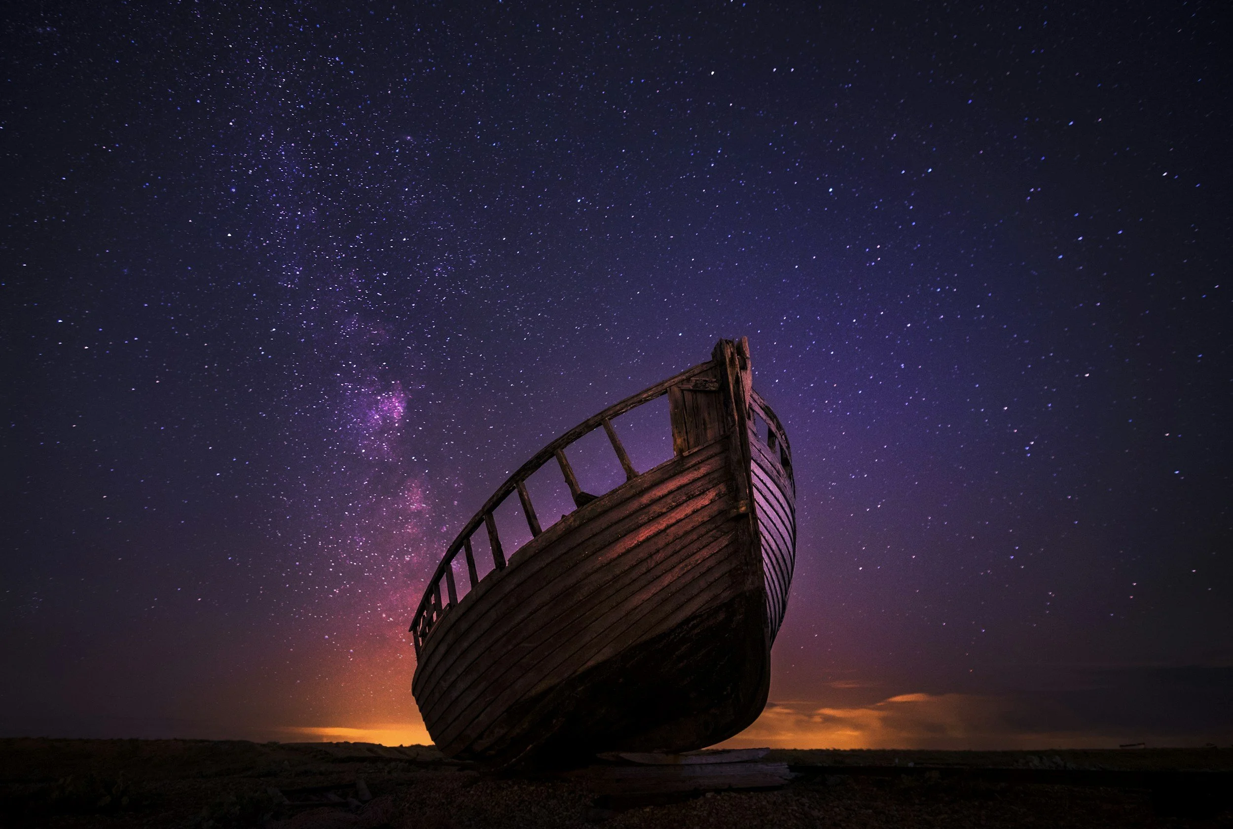 An old wooden shipwreck on a dark beach beneath a starry night sky with the Milky Way galaxy visible.
