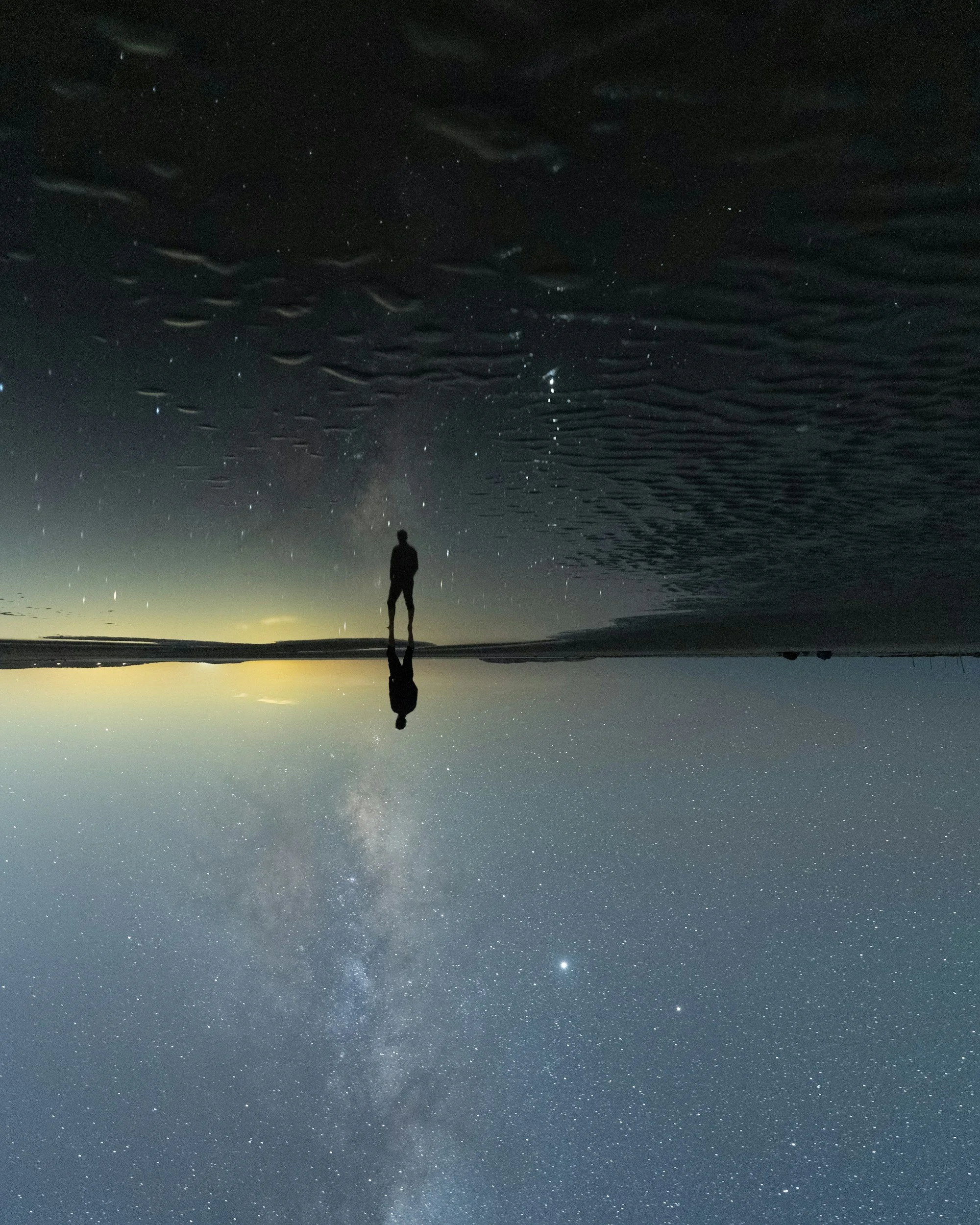 Silhouette of a person standing on the beach at night, with the starry sky and the Milky Way galaxy visible above, reflected in the water.